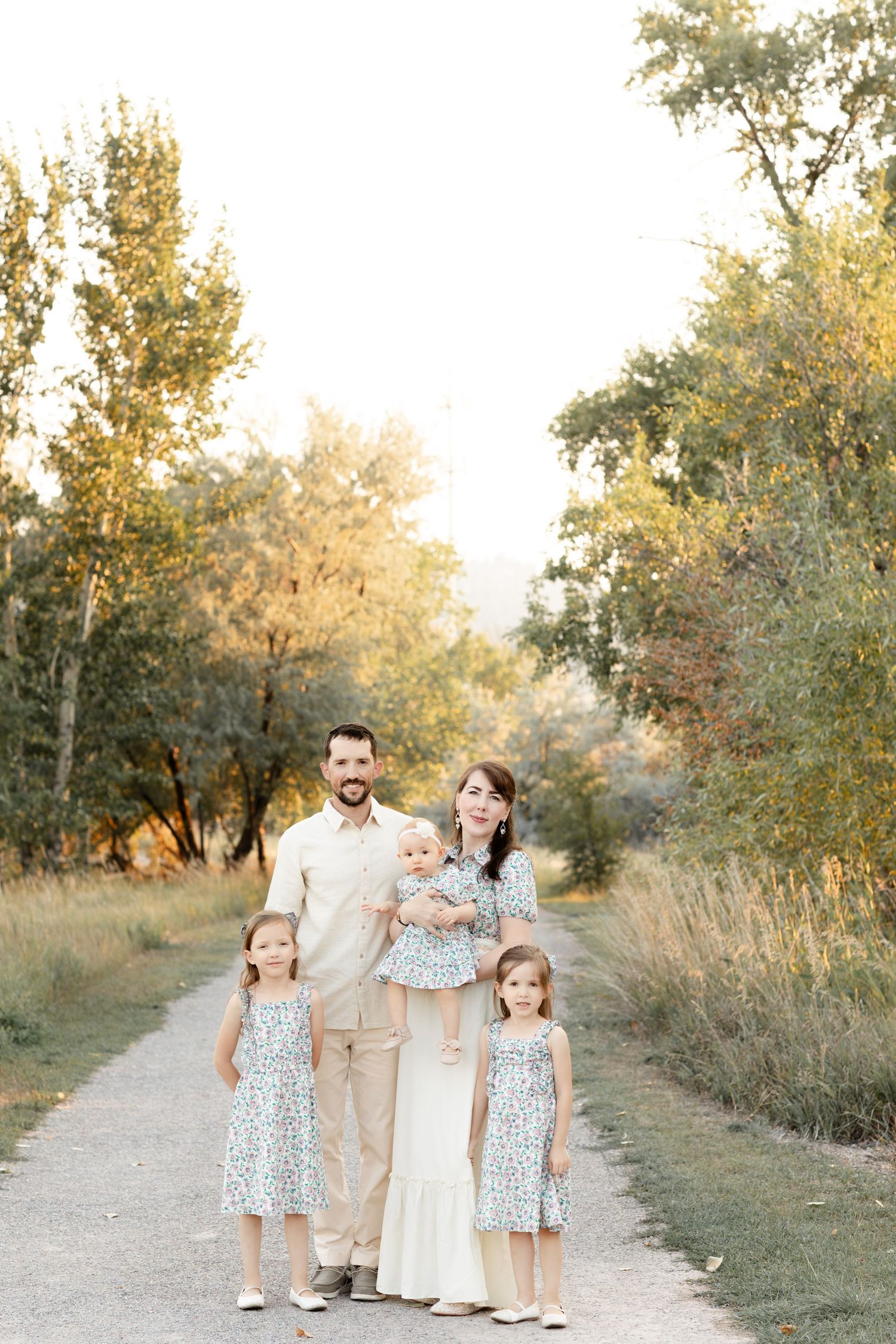 Family of five in neutral tones posing on tree-lined path during golden hour sunset.