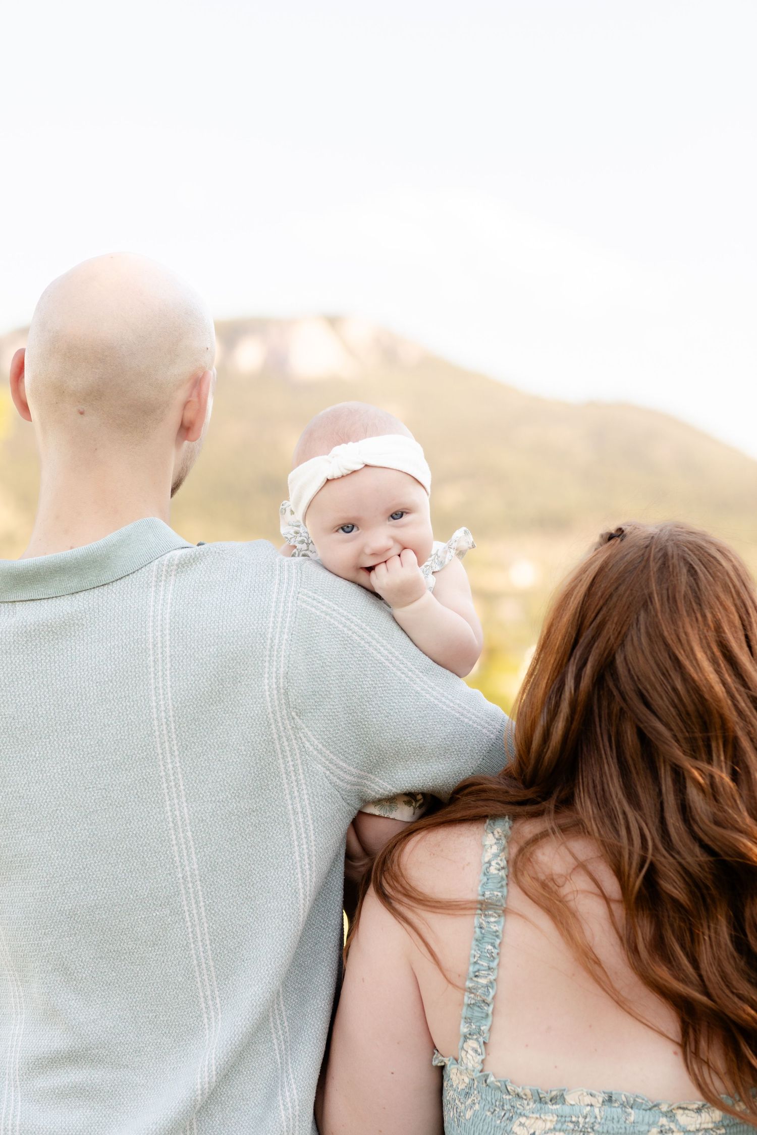 A bald father and redhead mother hold their smiling baby outdoors with mountains in the background.