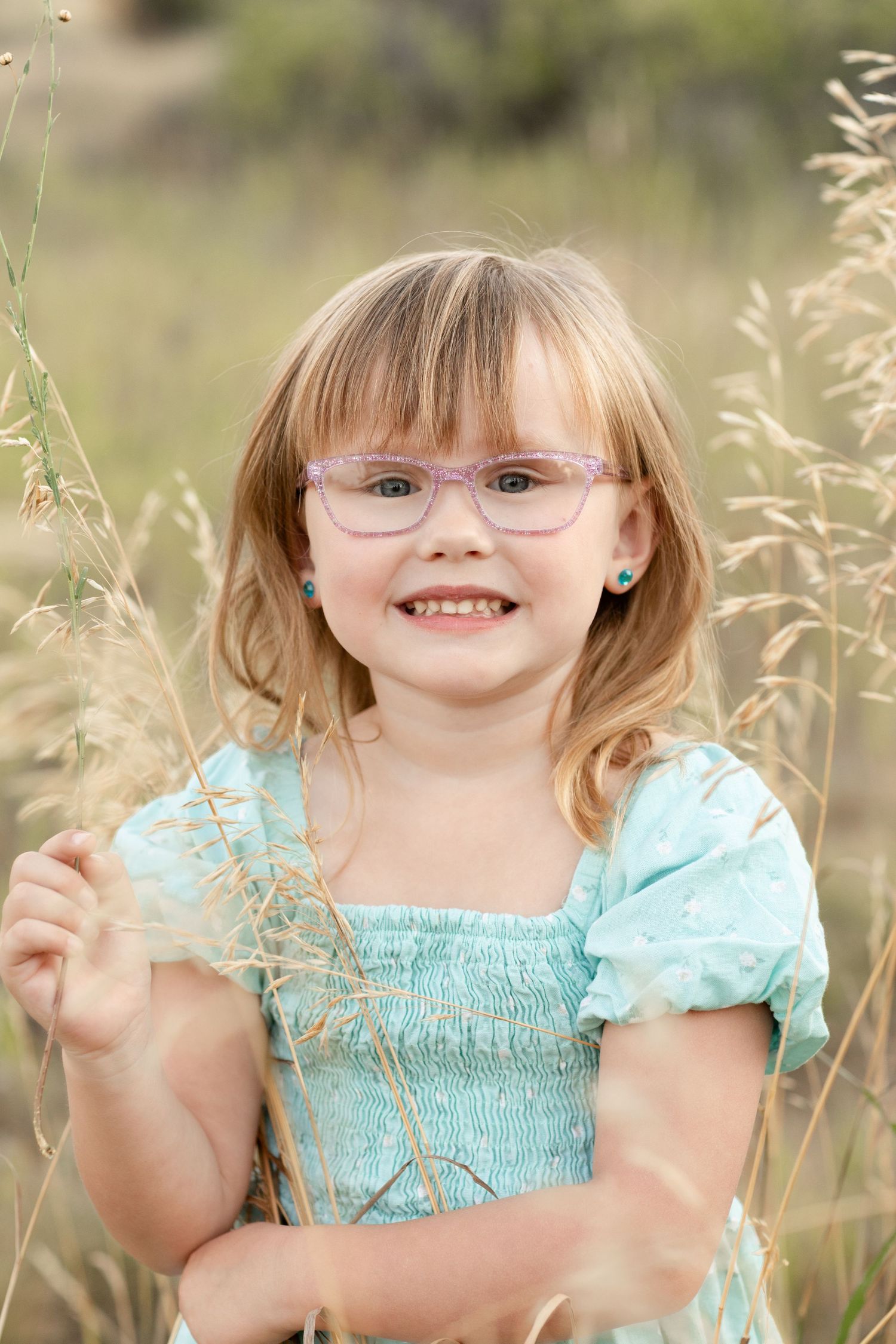 A smiling young girl with pink glasses and a teal dress stands among tall golden grasses in a field.