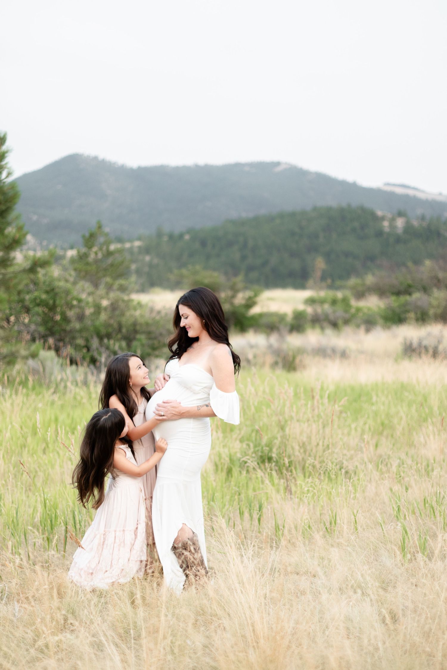 Pregnant mother in white dress with two daughters touching baby bump in meadow.