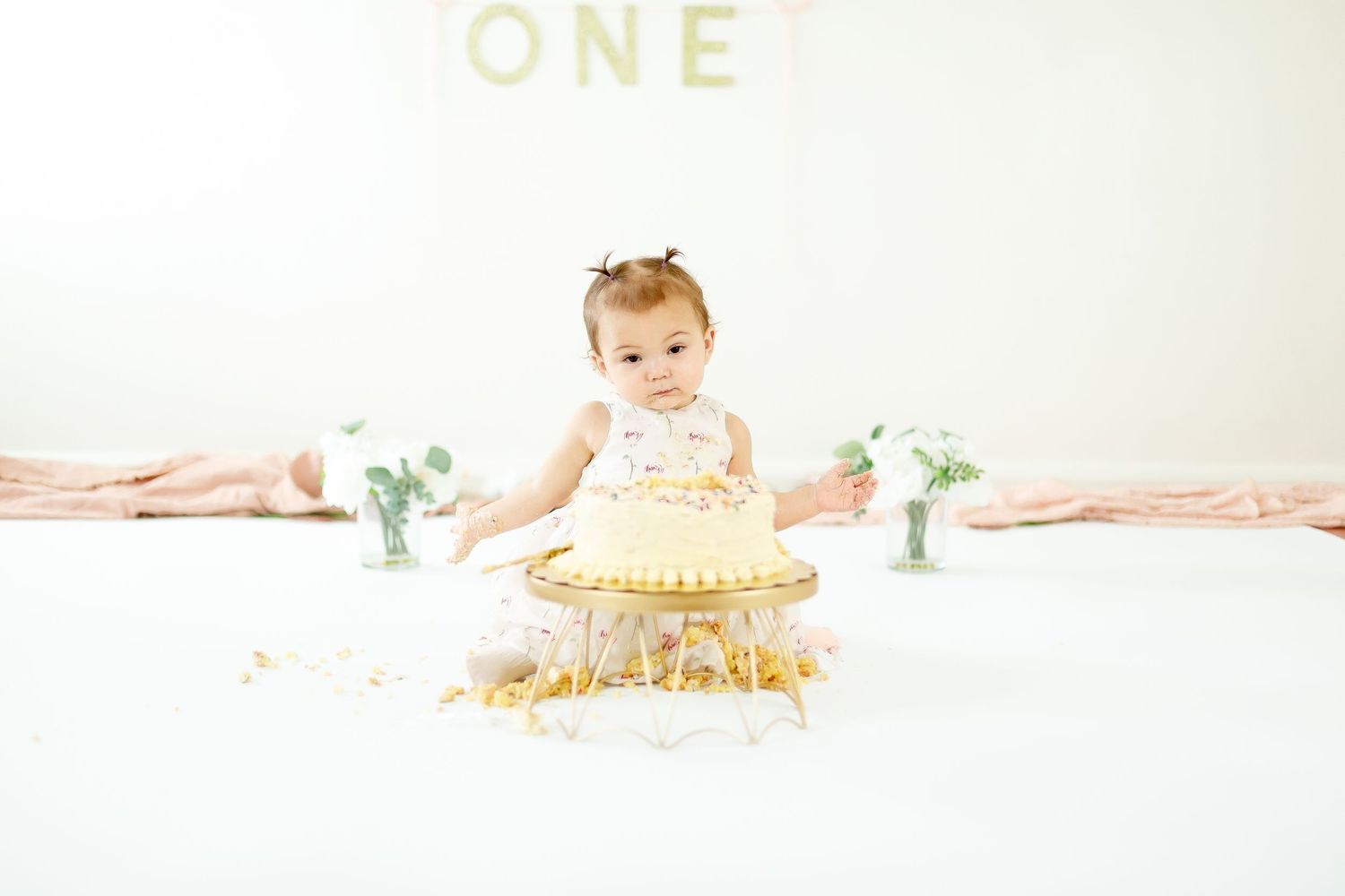 A one-year-old baby girl sits before a white smash cake during her first birthday photo session.