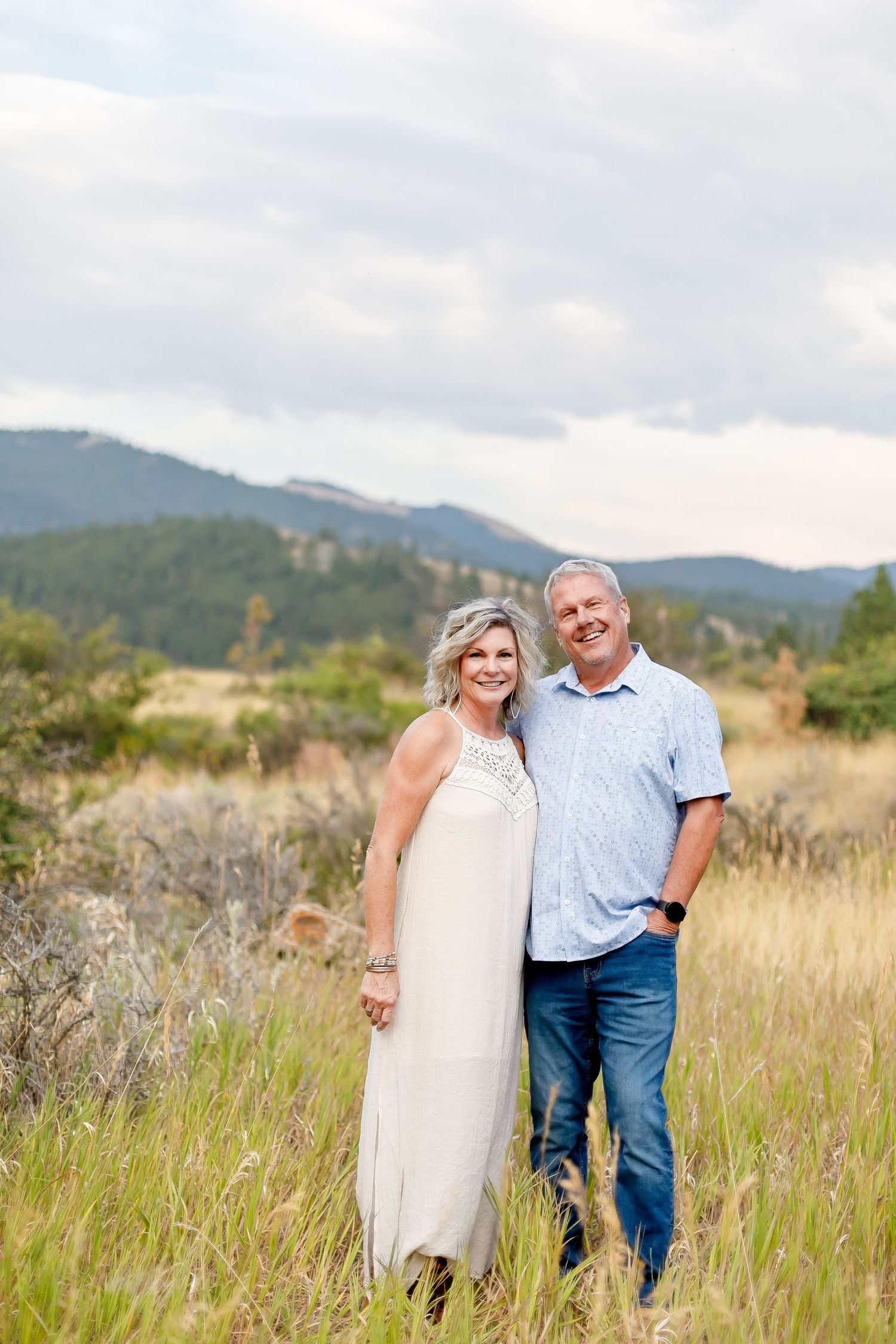 A mature couple posing in a mountain meadow, the woman in a white dress and man in a blue shirt.