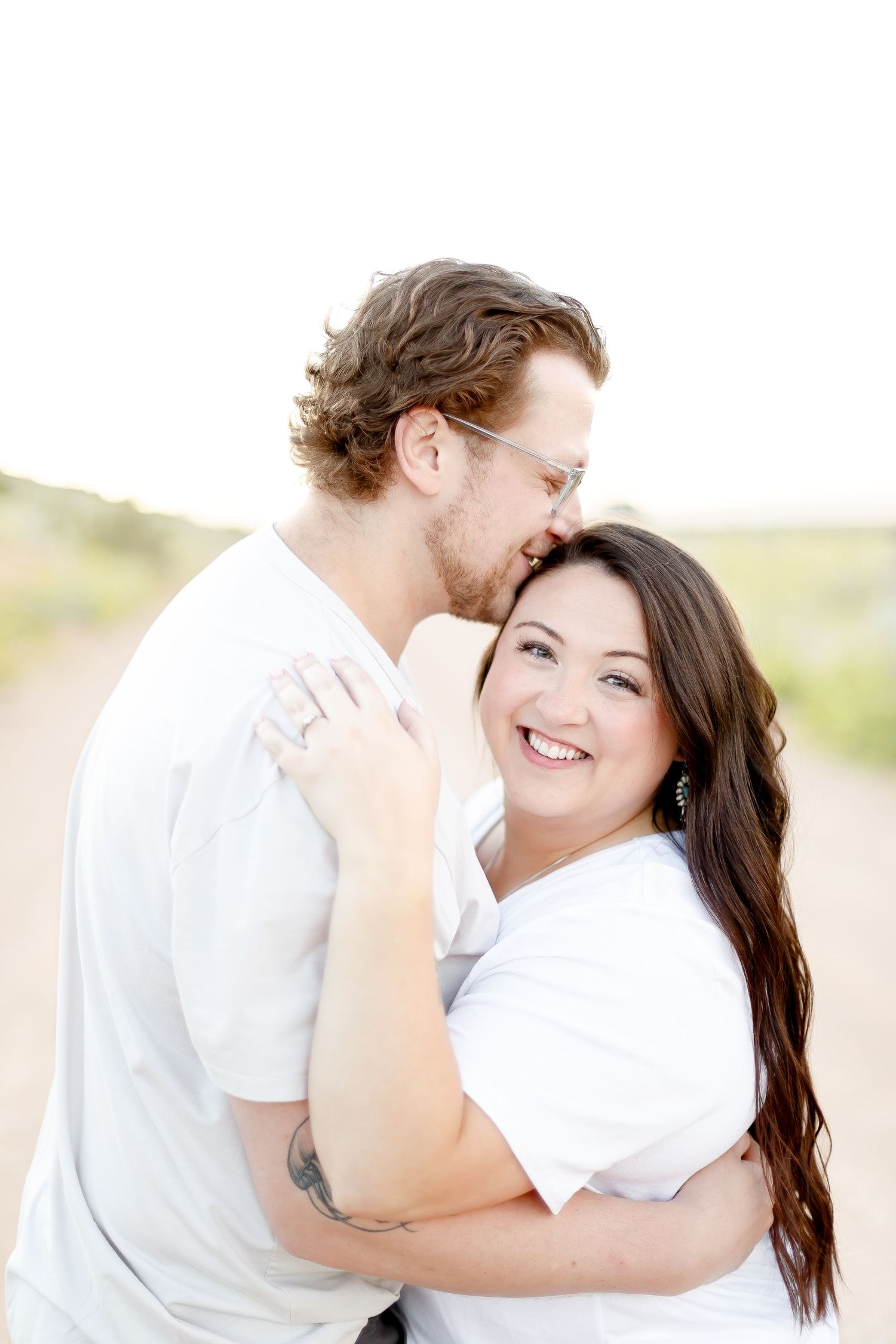 A joyful couple embracing outdoors, the man kissing her temple while she smiles at the camera.