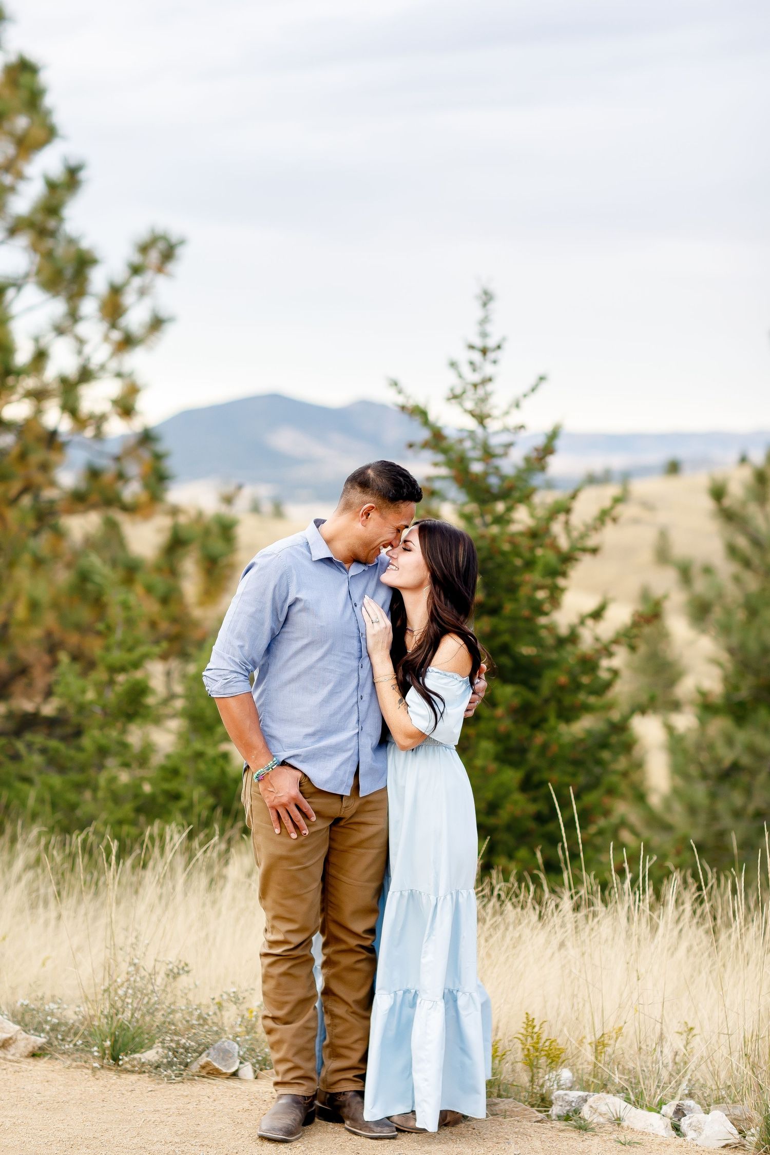 Couple embracing tenderly on a dirt path surrounded by pine trees and rolling hills.