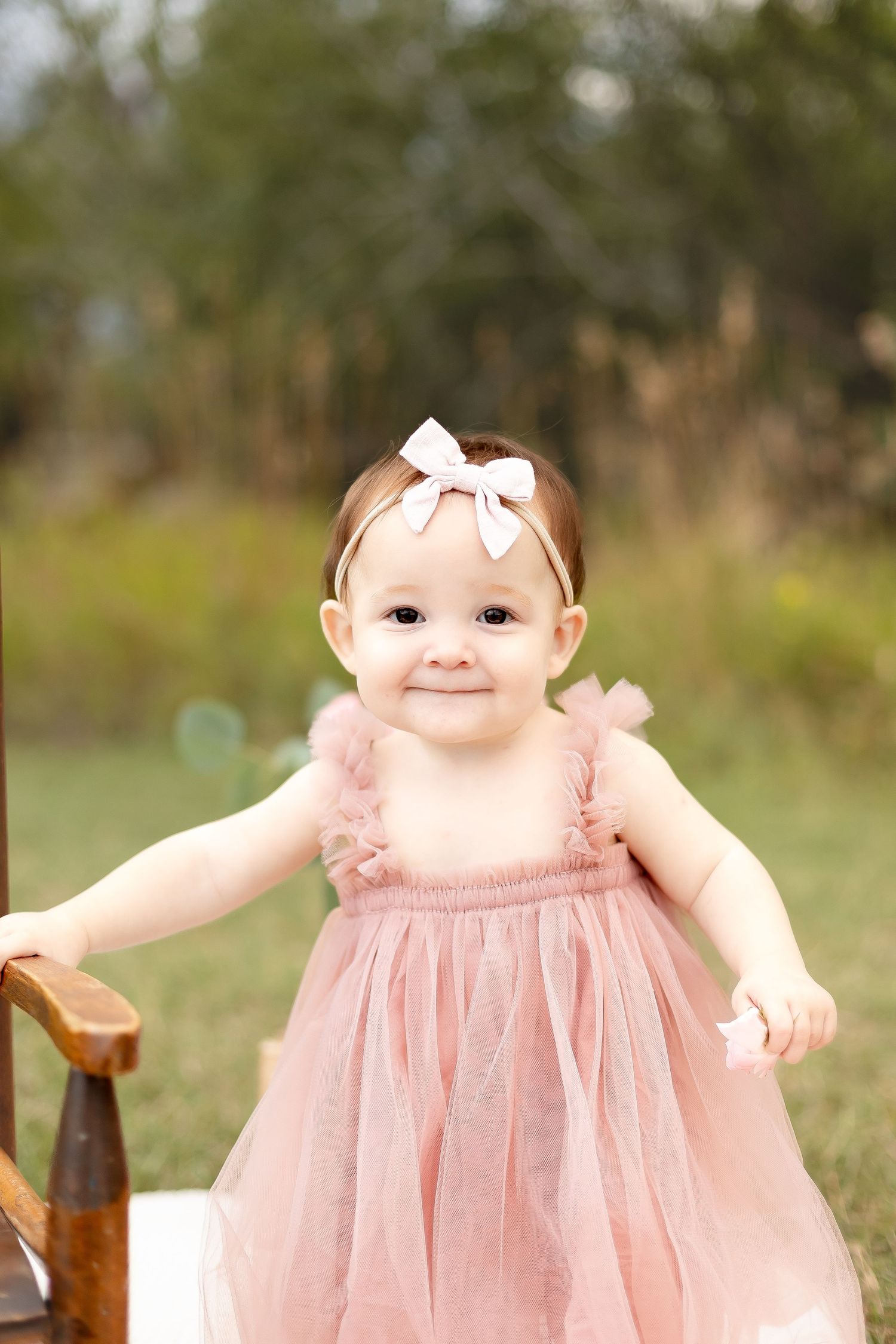 Adorable baby girl in pink tulle dress with white bow headband smiles during outdoor birthday photo session.