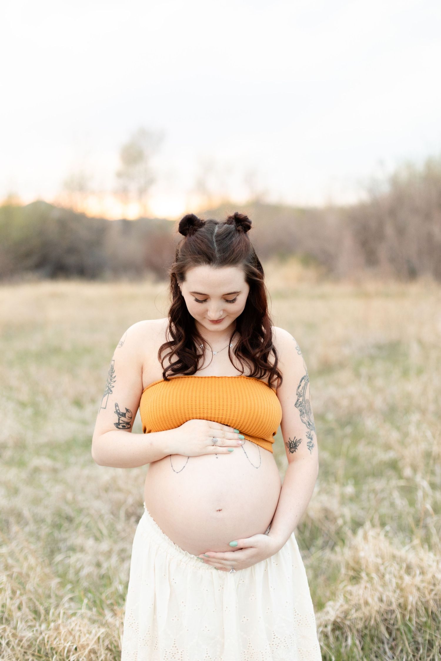 Pregnant woman in orange crop top and white skirt cradles her bare baby bump in an open field at dusk.