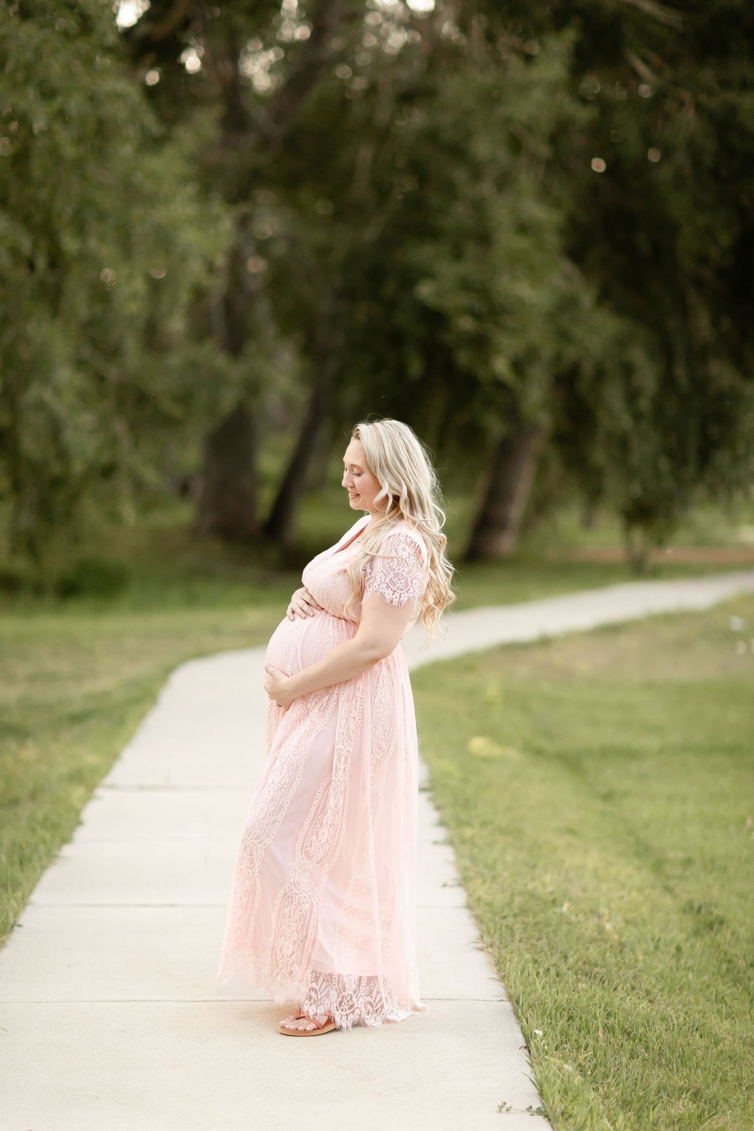 Pregnant blonde woman in pink lace maxi dress standing on garden path surrounded by trees.