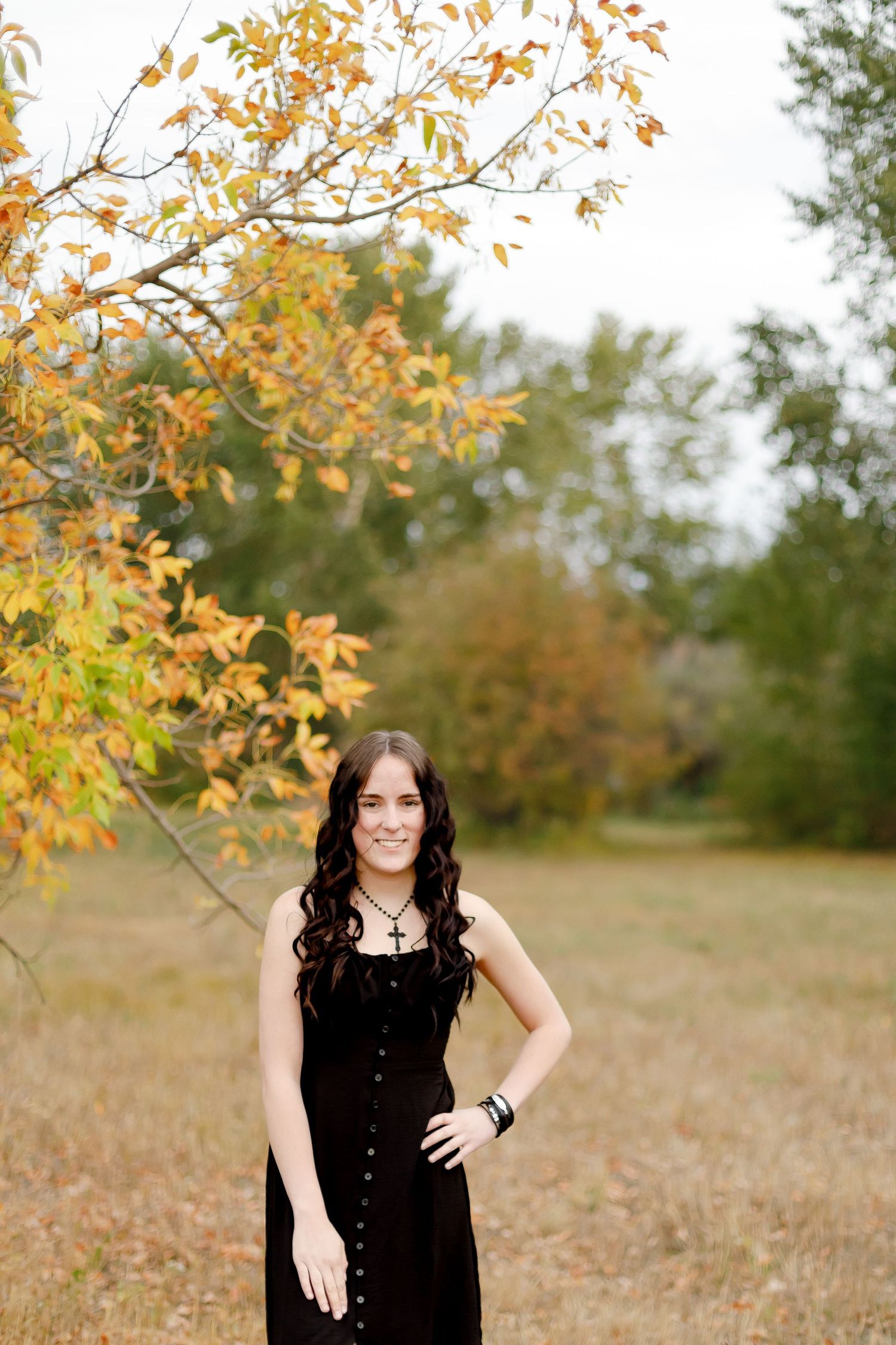 Smiling young woman in black dress standing in a field under colorful autumn foliage.