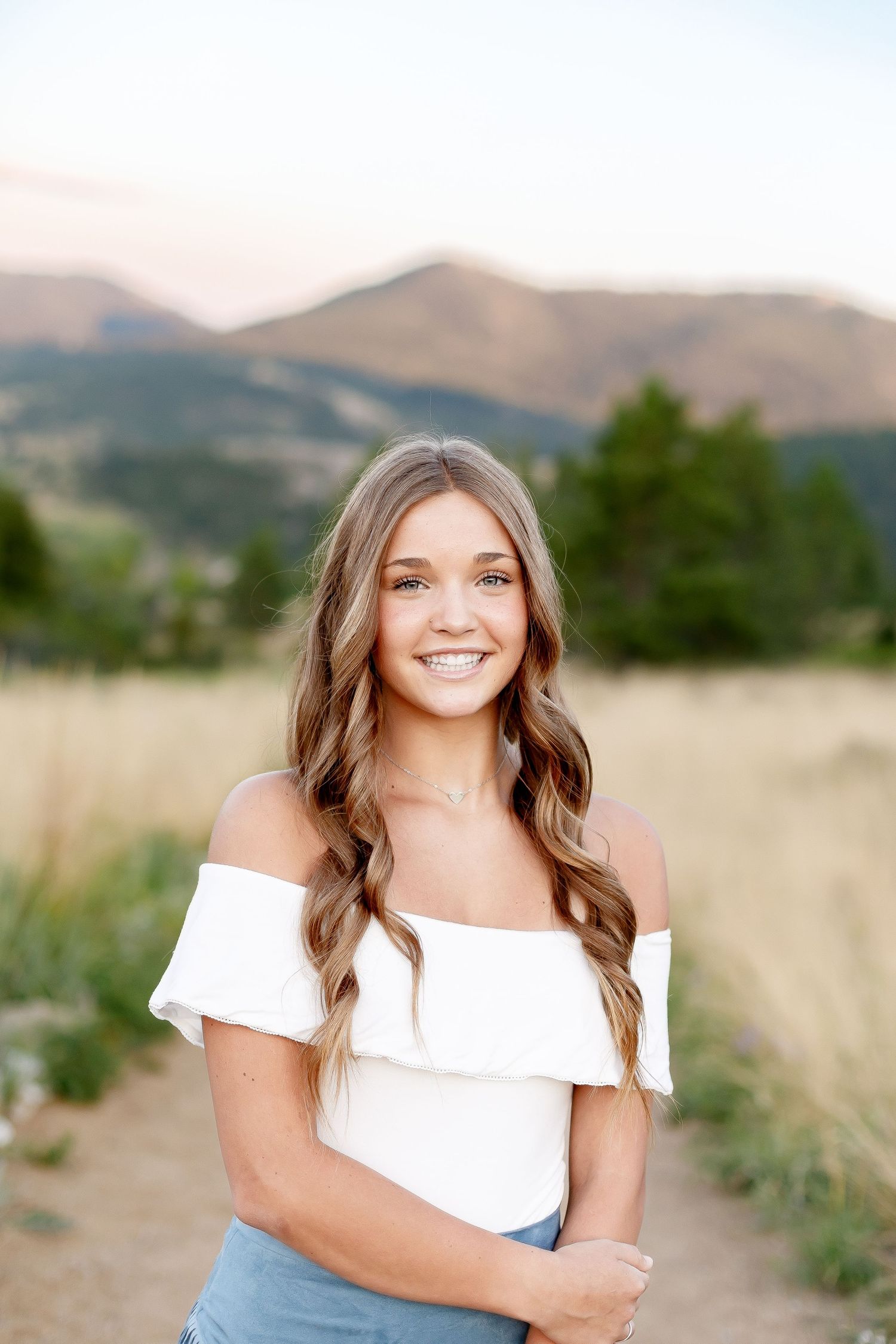 Smiling young woman in white off-shoulder top with mountain landscape backdrop at golden hour.