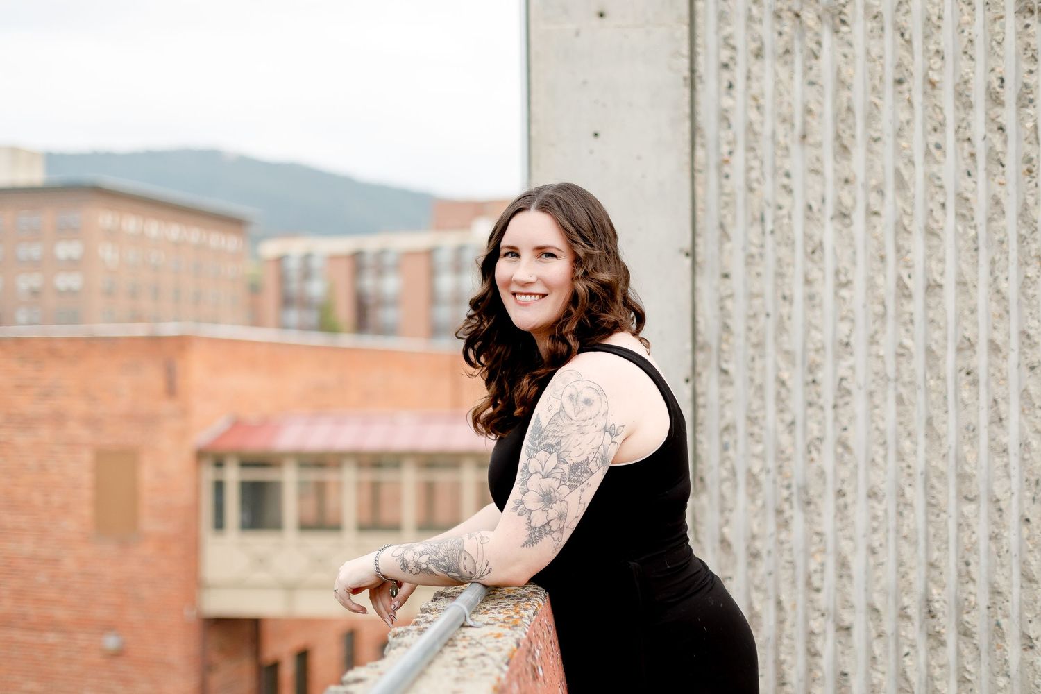 Smiling tattooed woman in black dress leaning on a railing with brick buildings in the background.