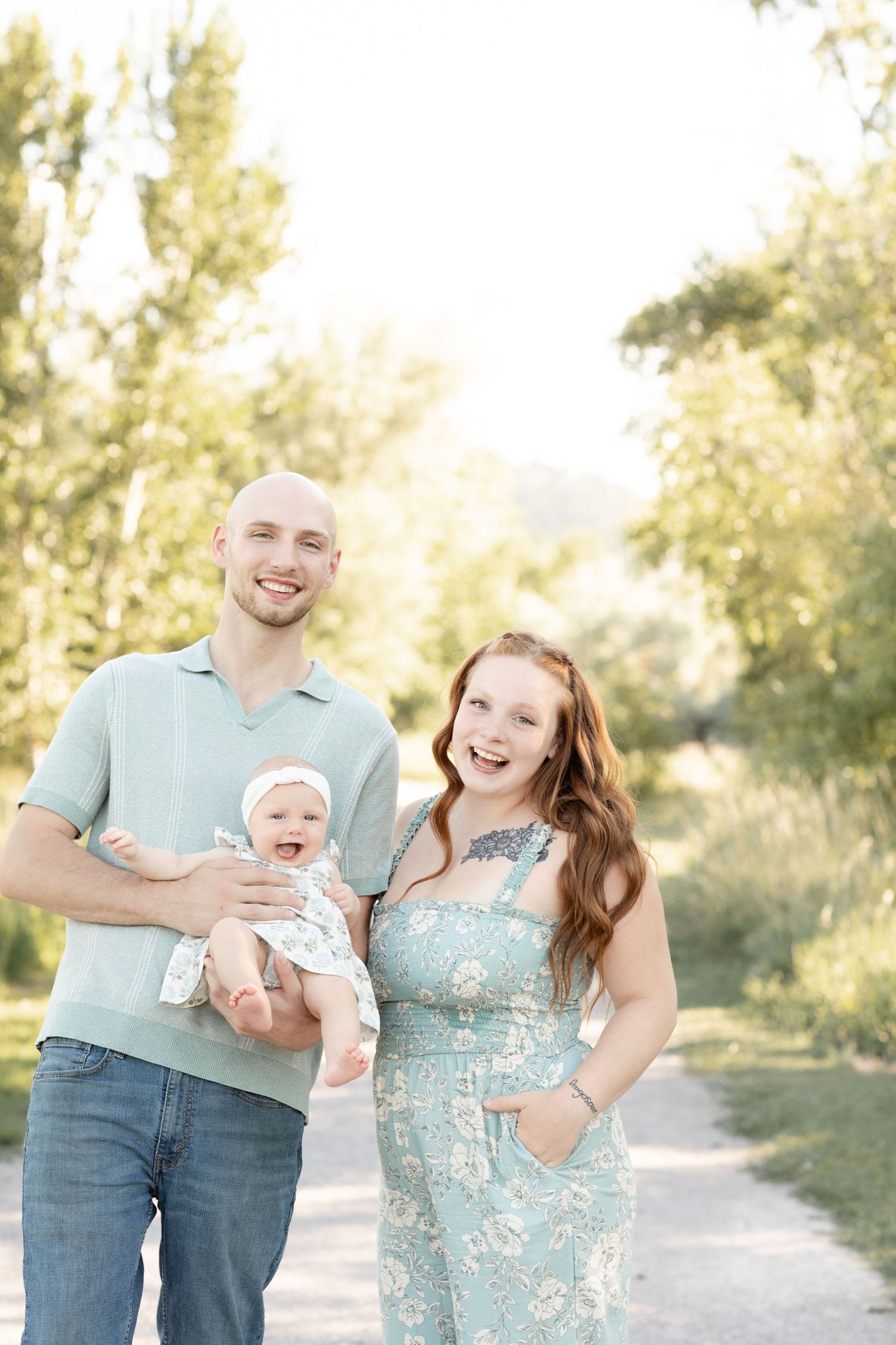 Young family with bald father, redhead mother in floral dress, and baby girl posing on sunny park path.
