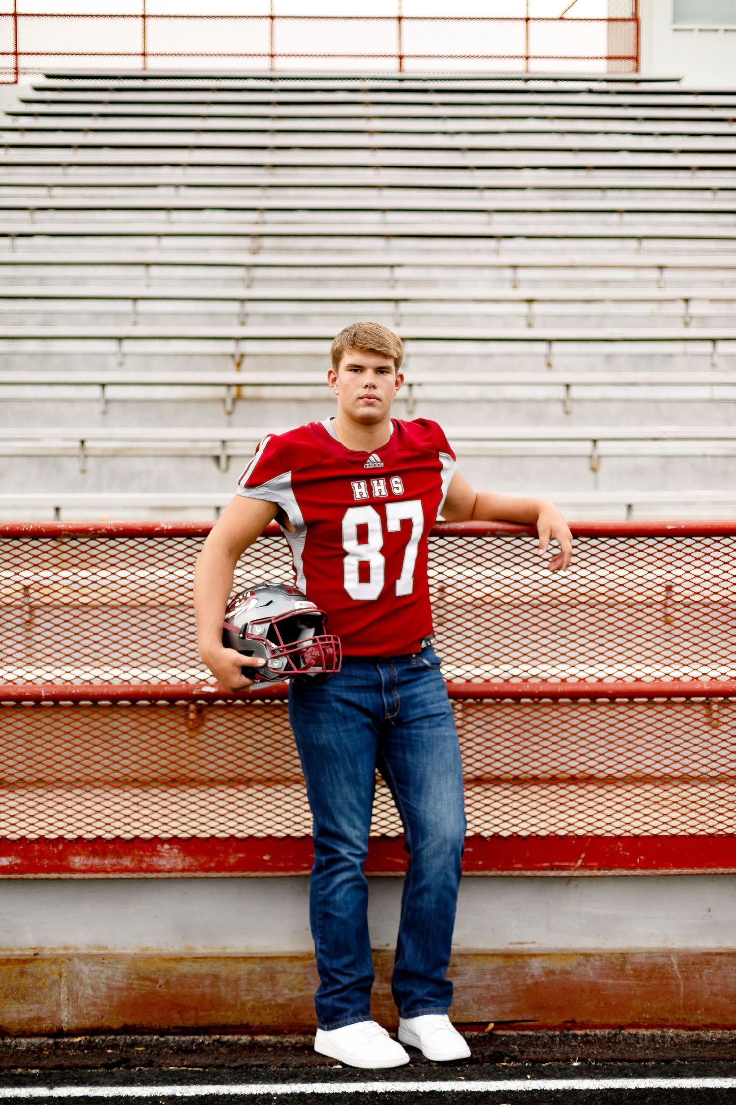Teen in HHS #87 red football jersey holding helmet while standing on stadium bleacher steps.