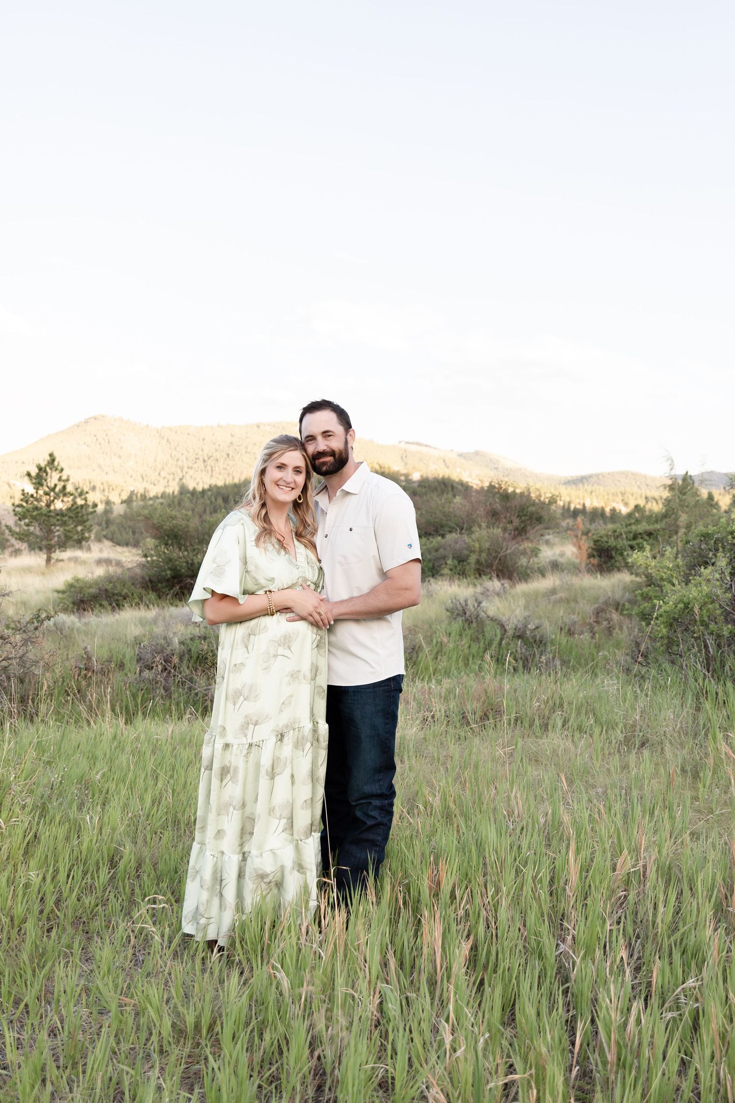 Expecting couple embracing and smiling together in lush green field with mountain views.