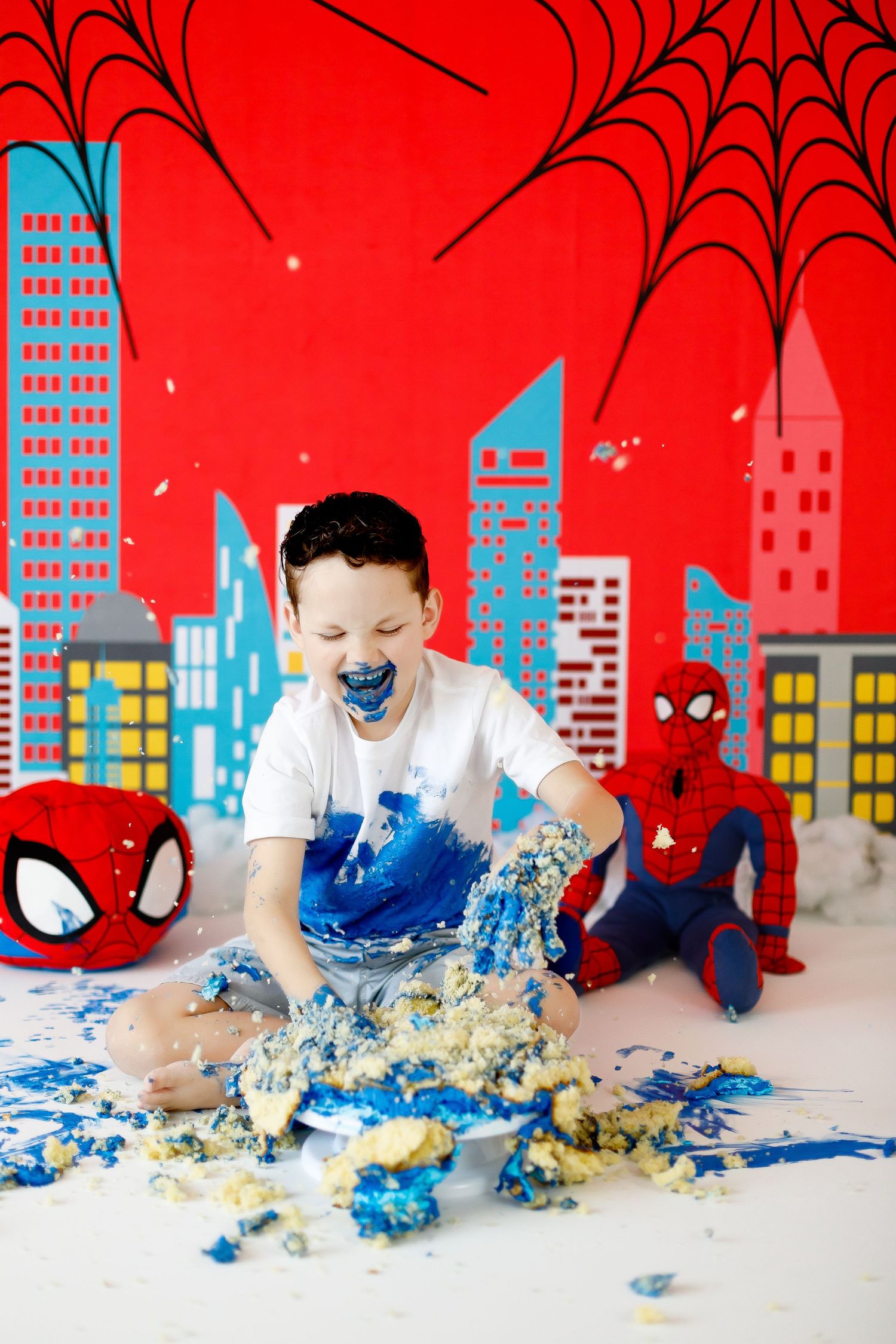 A boy laughs while smashing a blue birthday cake surrounded by Spiderman toys and decor.