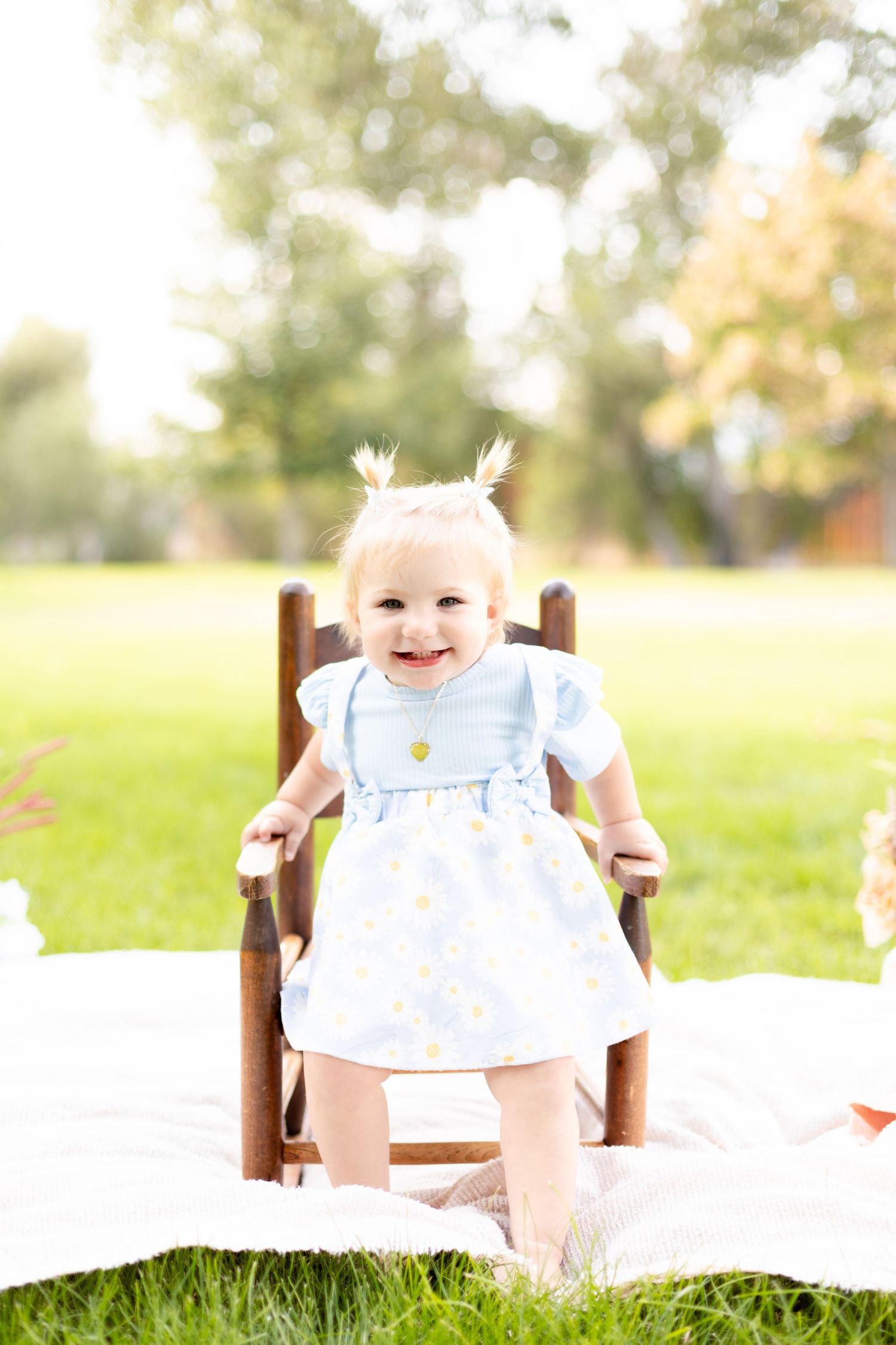 Smiling baby girl in a blue daisy dress standing in a wooden chair outdoors in a sunny green park.