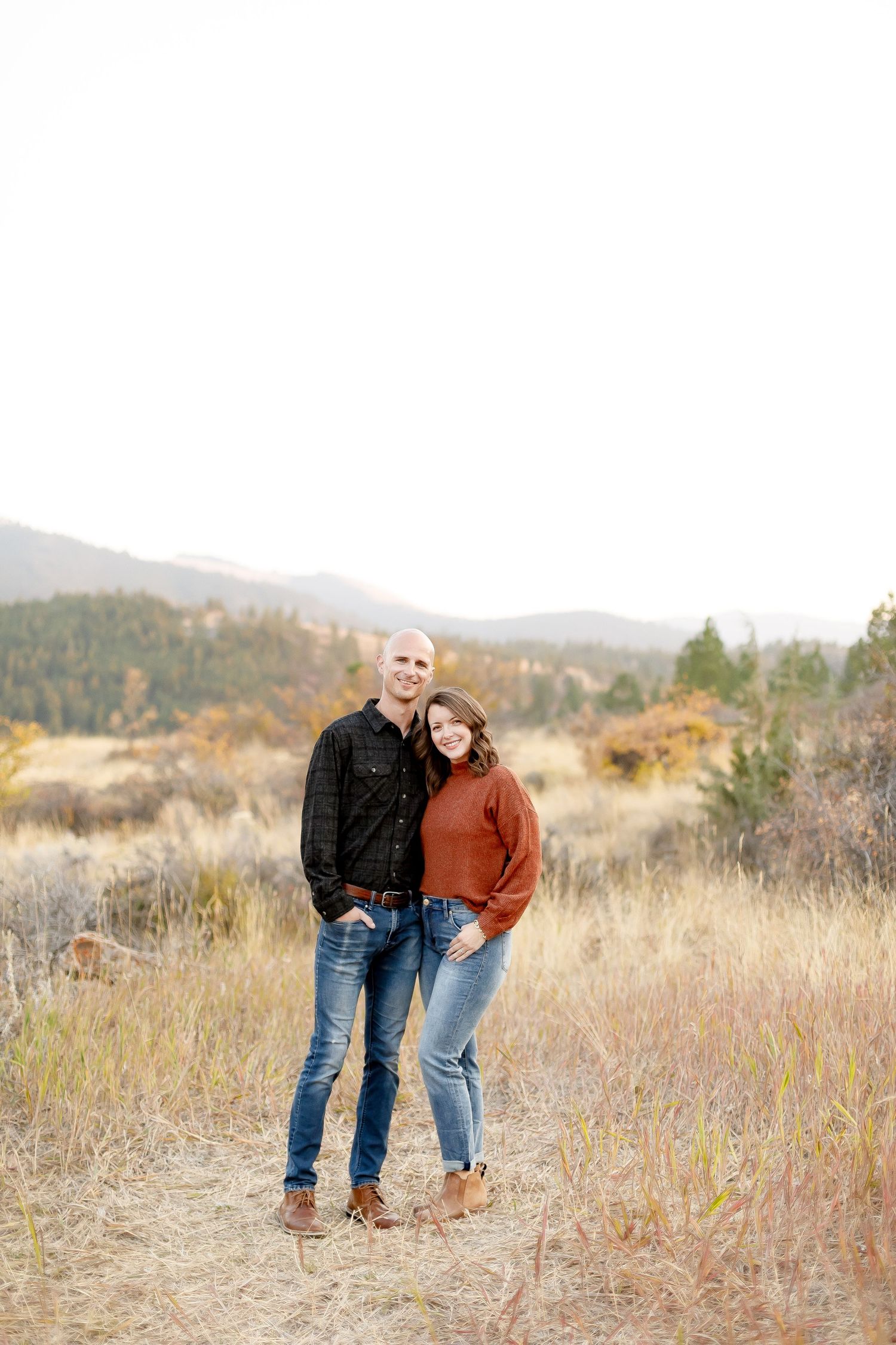 Smiling couple posing together on a trail surrounded by fall foliage and mountain scenery.