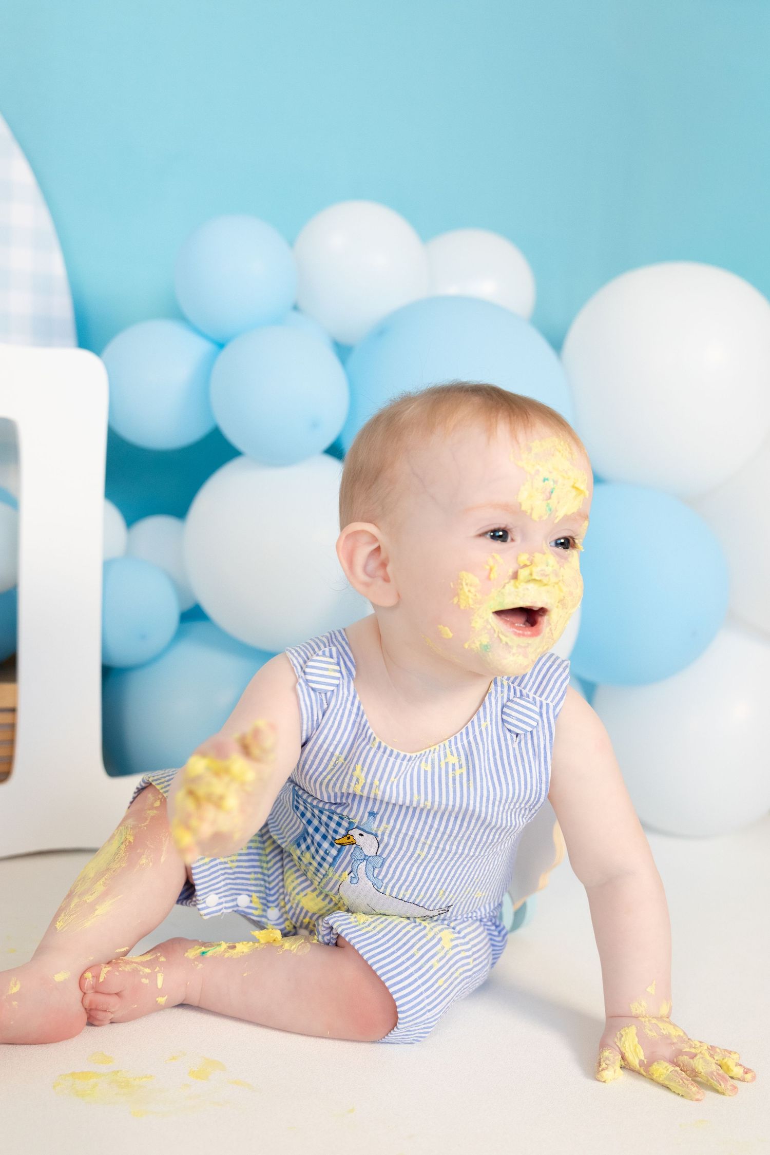 Happy baby covered in yellow cake frosting laughing during blue balloon cake smash photoshoot.