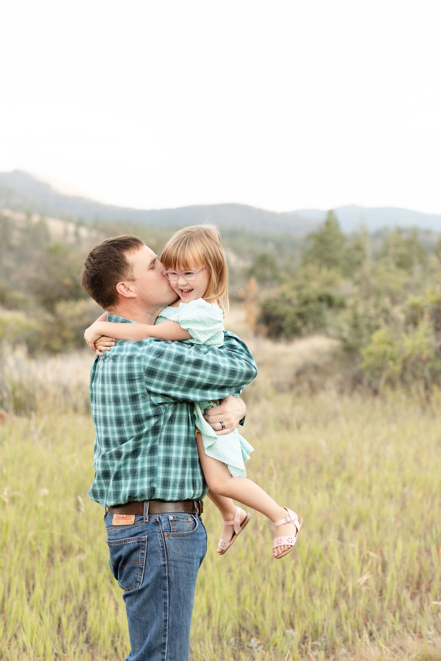 A father kisses his laughing young daughter while holding her in a scenic outdoor field.