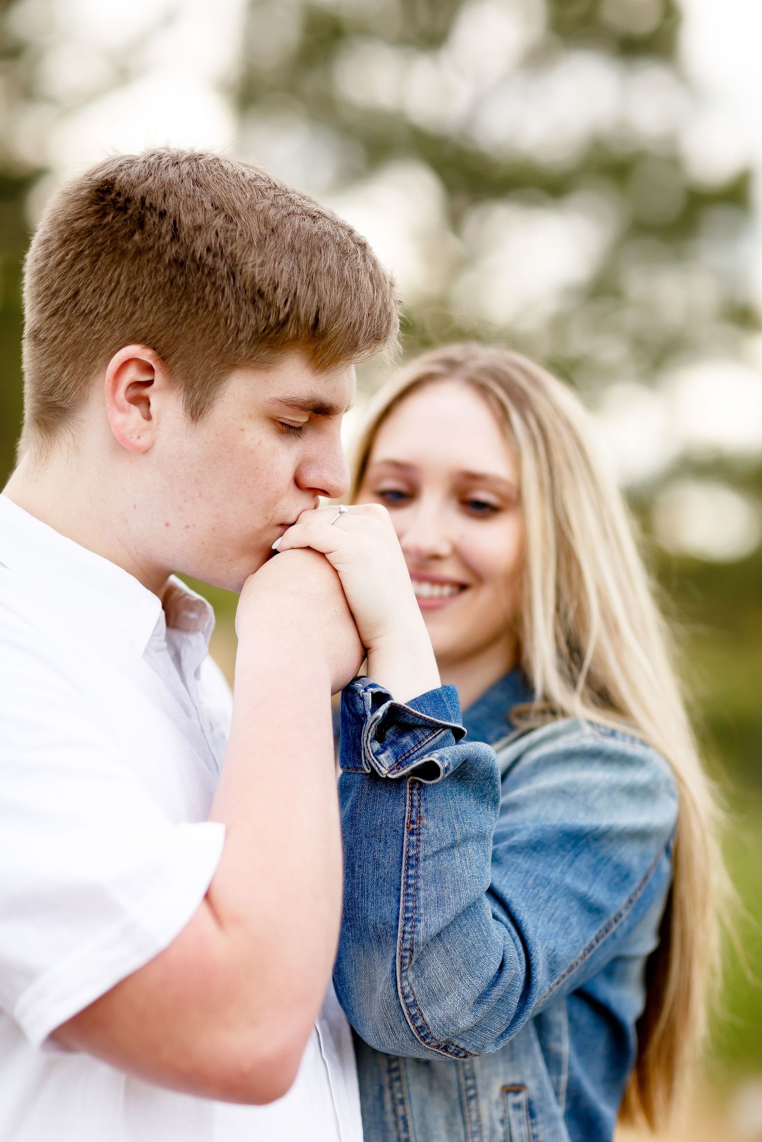 Young man kissing his fiances hand while she smiles during an outdoor engagement session.