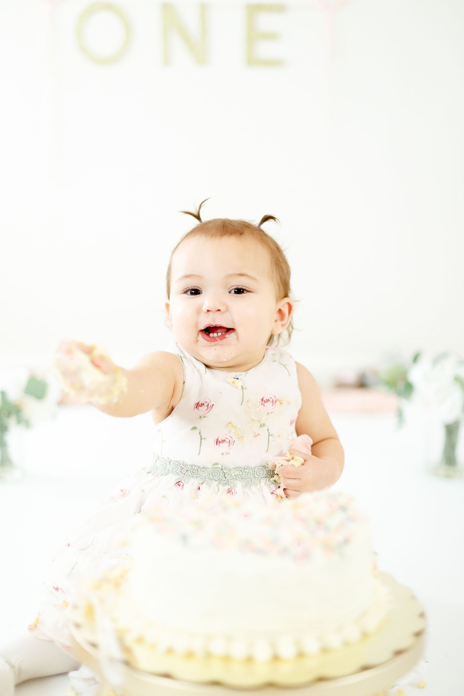 A joyful baby girl reaches toward her first birthday smash cake with a happy open-mouthed smile.