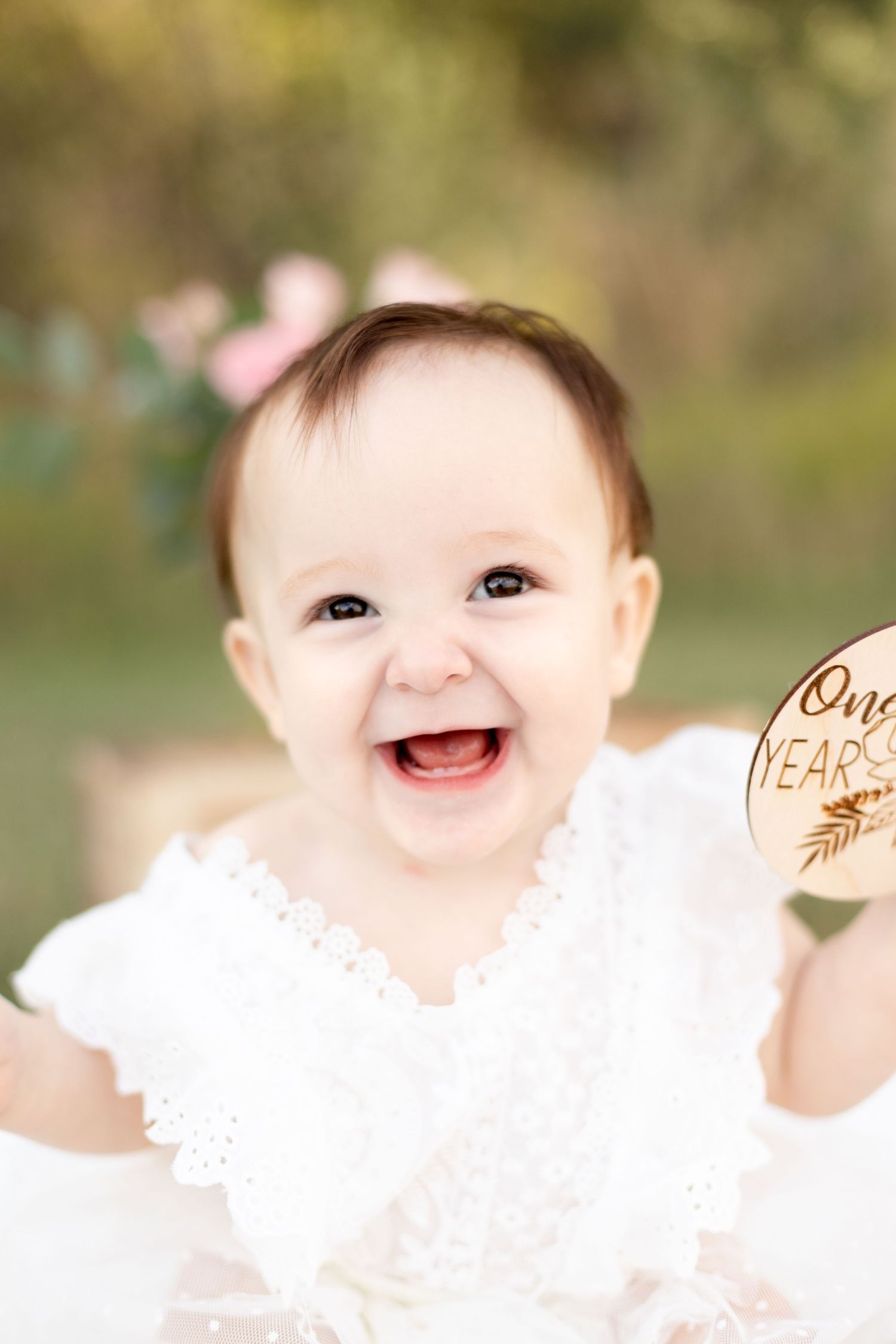 Happy baby girl laughing while holding a wooden One Year milestone sign during birthday photoshoot.
