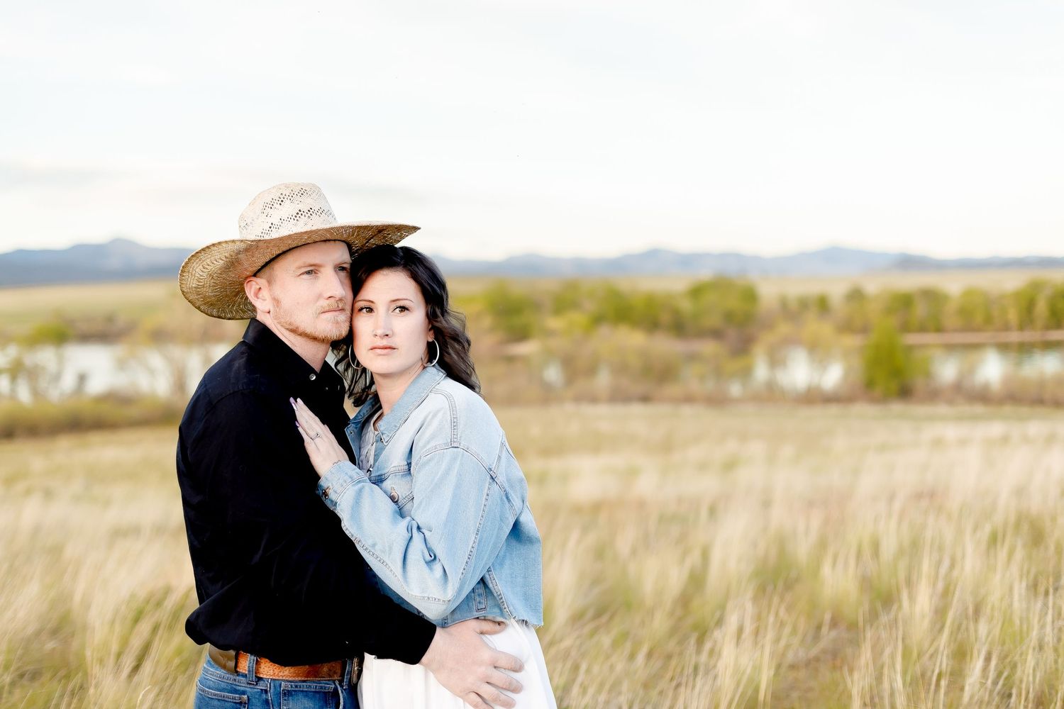 Couple embracing in golden field with mountains, man in cowboy hat and black shirt, woman in denim jacket.