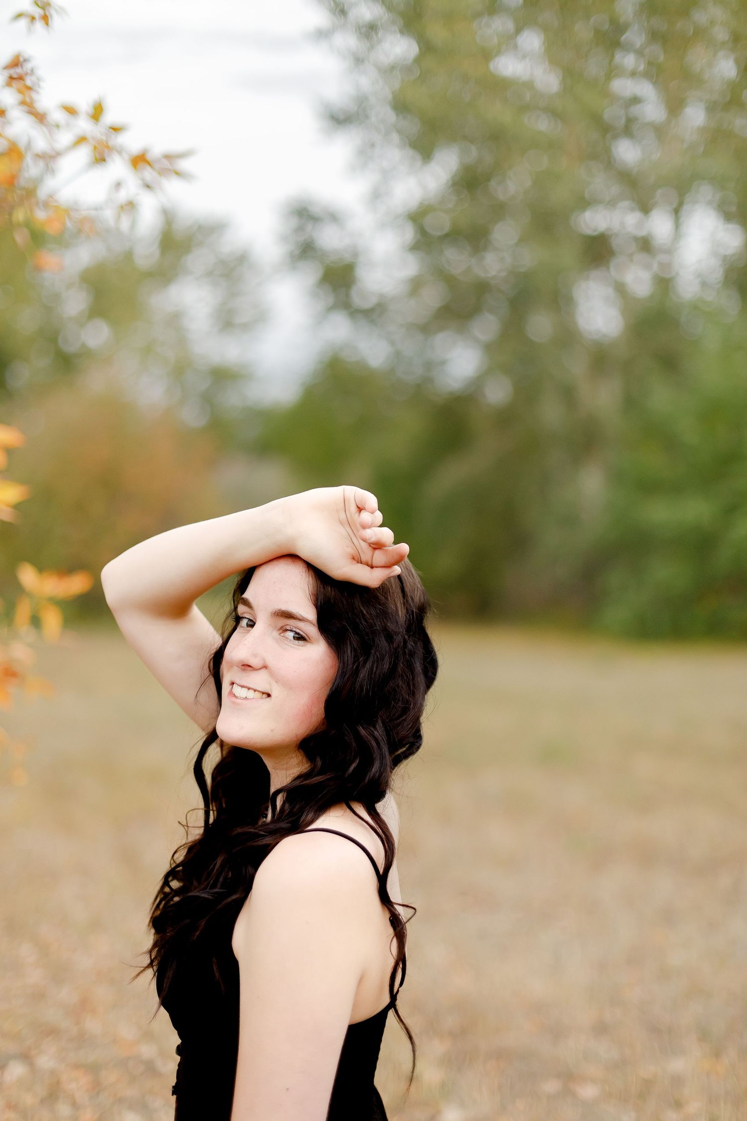 Young woman with dark curly hair smiling outdoors in a black dress, posing in a natural park setting during autumn.