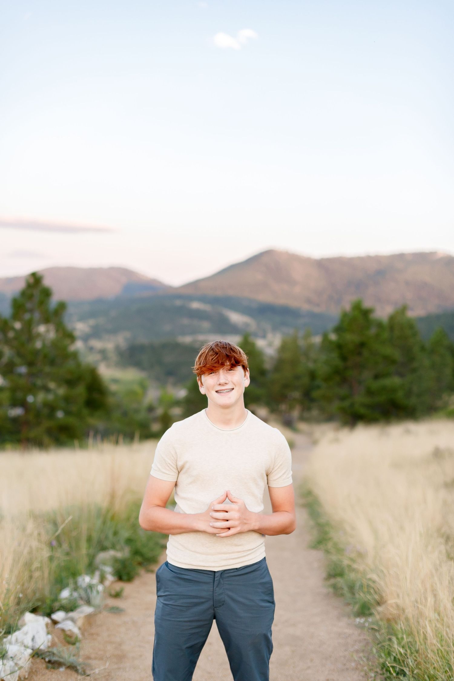 Cheerful young man with red hair in beige t-shirt posing on a scenic mountain path at sunset.