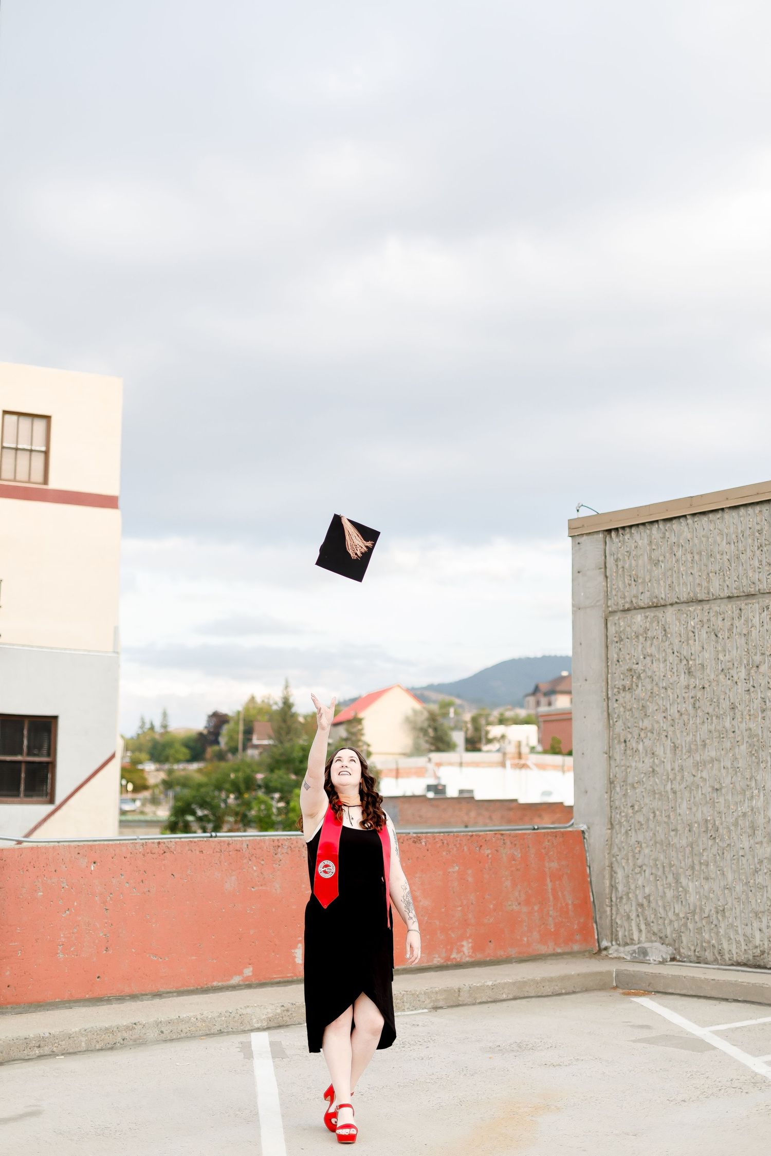 Graduate in black dress tossing mortarboard cap in the air on a rooftop with city skyline behind her.