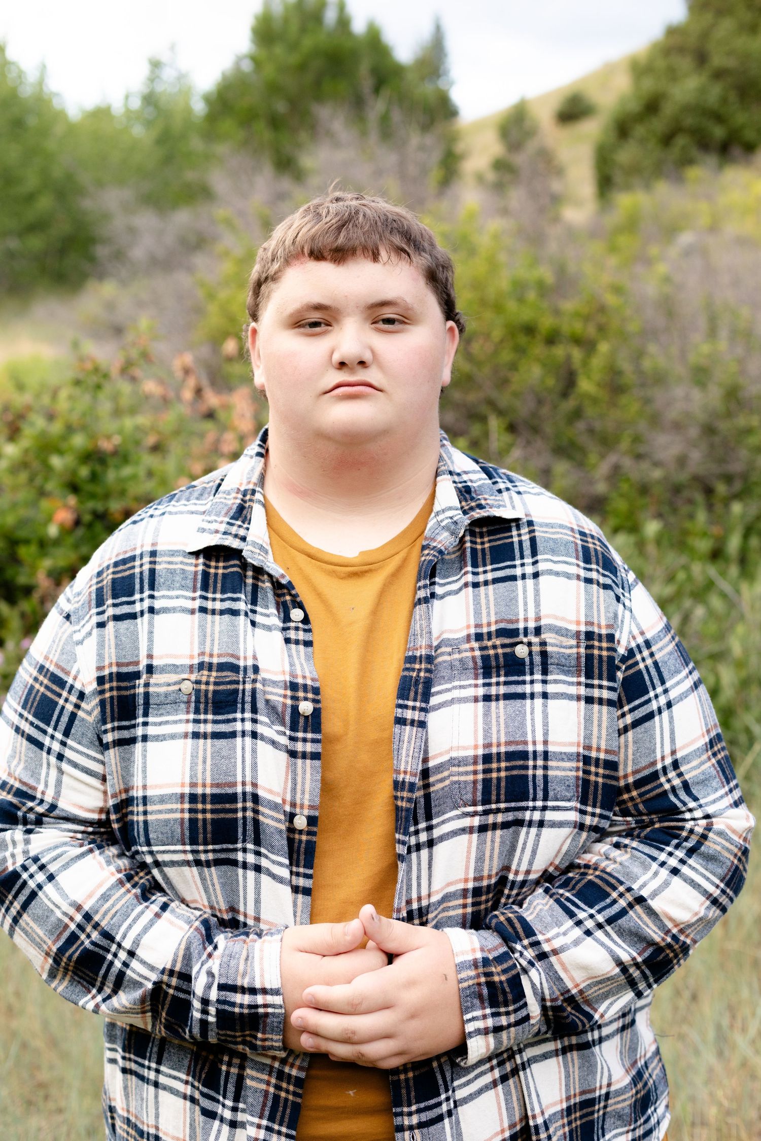 Young person in plaid flannel shirt standing confidently in a natural outdoor setting with green foliage background.