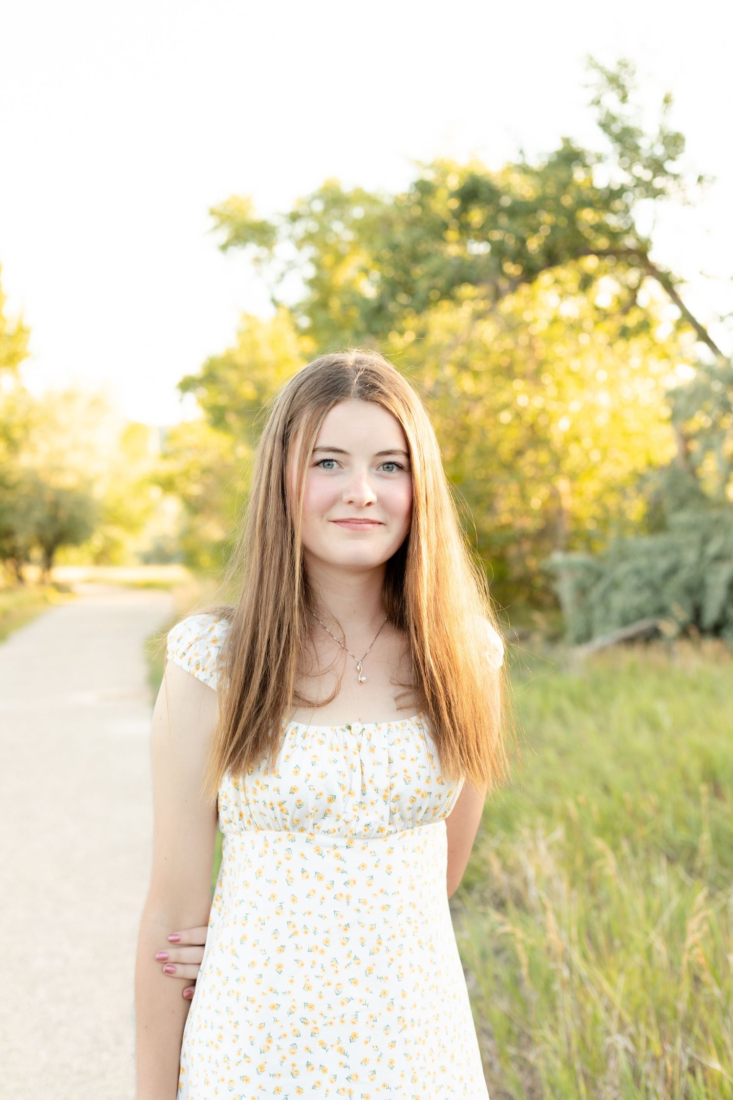 Young woman with long blonde hair in a floral sundress standing on a sunlit path surrounded by trees.