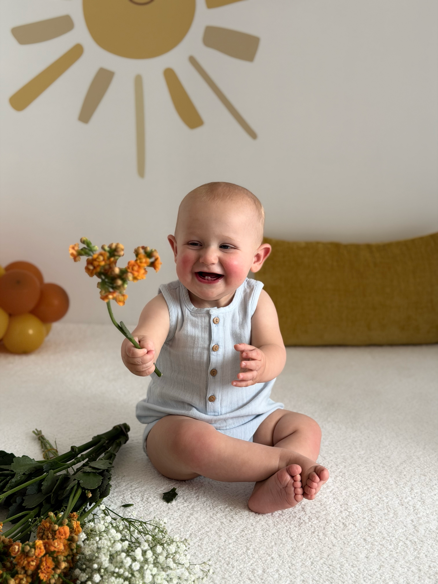 Happy laughing baby in blue romper holding orange flowers with sun wall art backdrop.