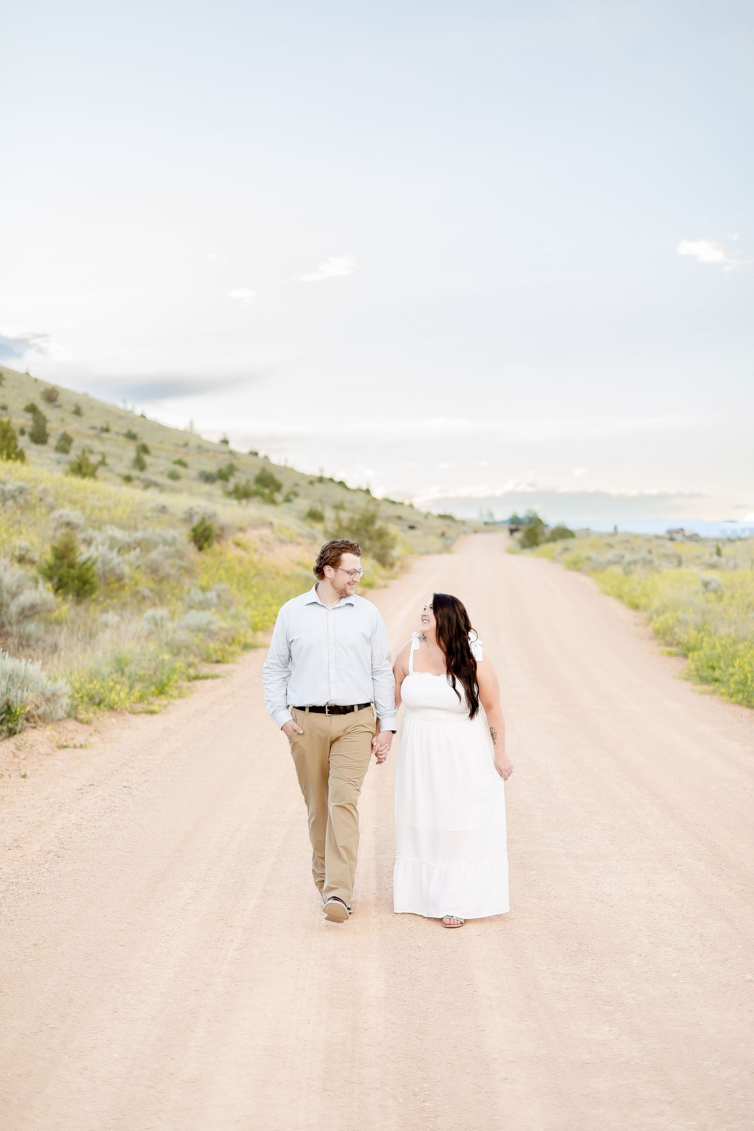 Couple walking hand in hand down a dirt road surrounded by hills, woman in white dress, man in khakis.