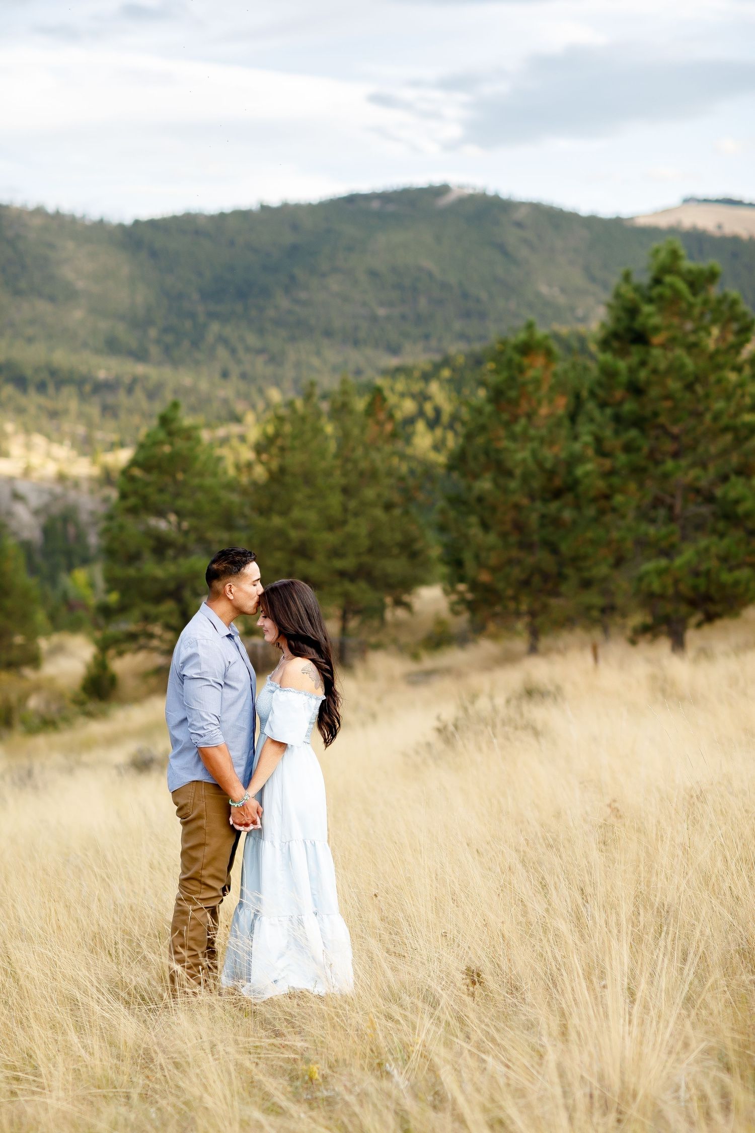 Couple sharing a romantic kiss while holding hands in tall golden grass with a scenic mountain backdrop.
