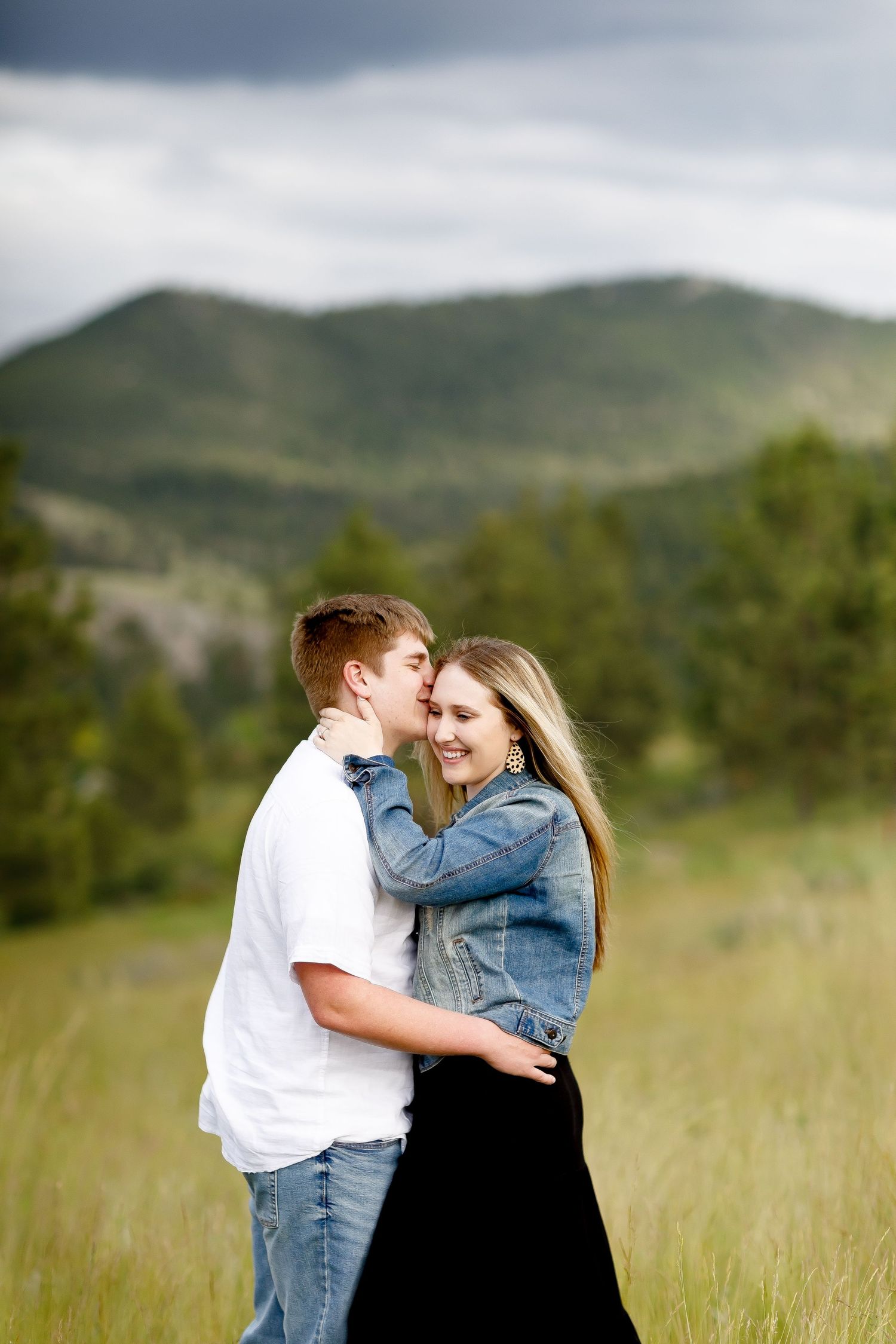 Young couple sharing a kiss in a mountain meadow with dramatic cloudy sky backdrop.