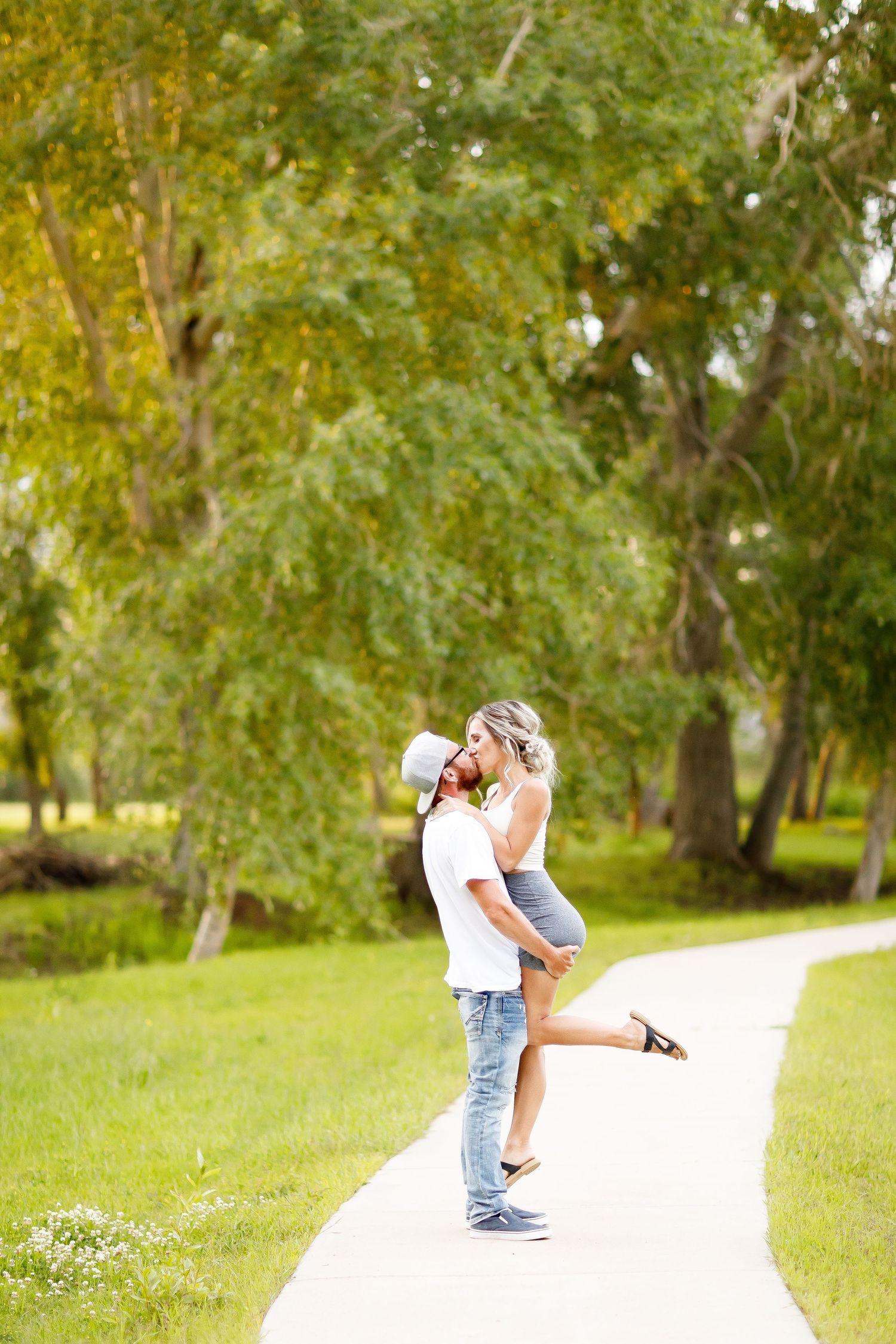 Man lifting woman for a romantic kiss on a tree-lined path during an outdoor couples engagement session.