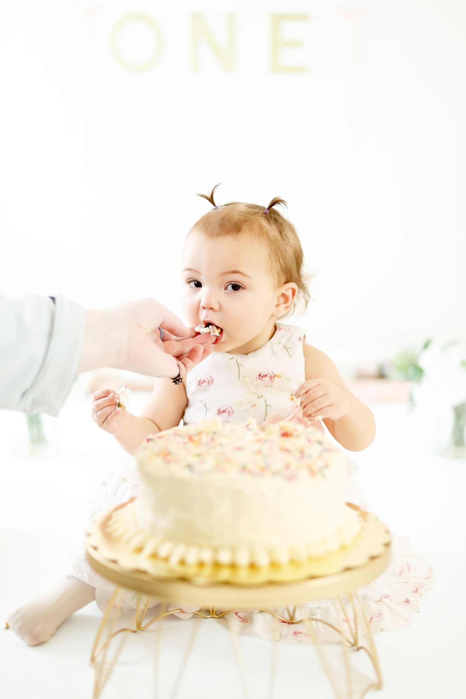 Baby girl in floral dress eating first birthday white cake being fed by adult hand.