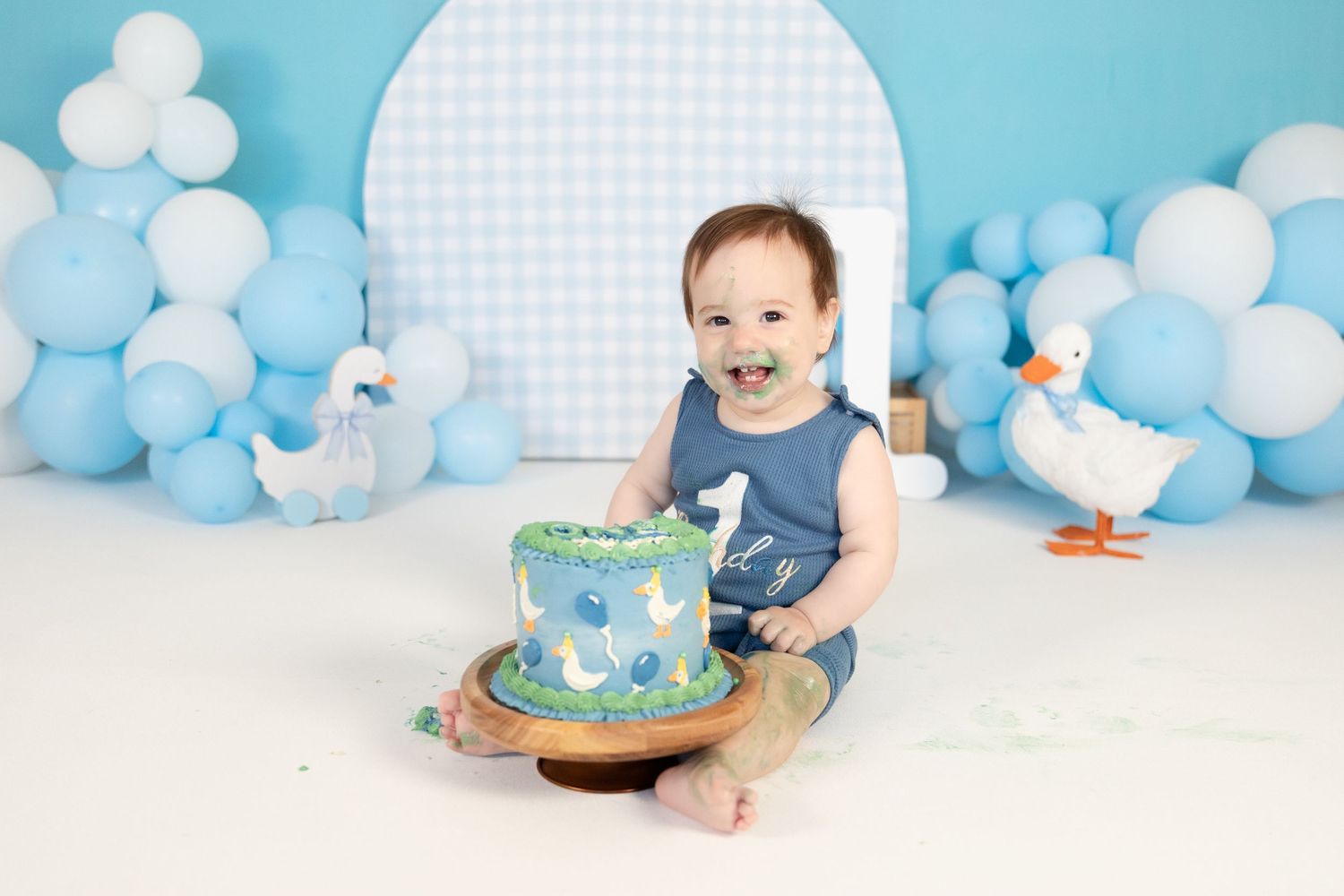 Smiling baby boy in blue romper holds duck-themed first birthday cake with balloon and duck decorations.