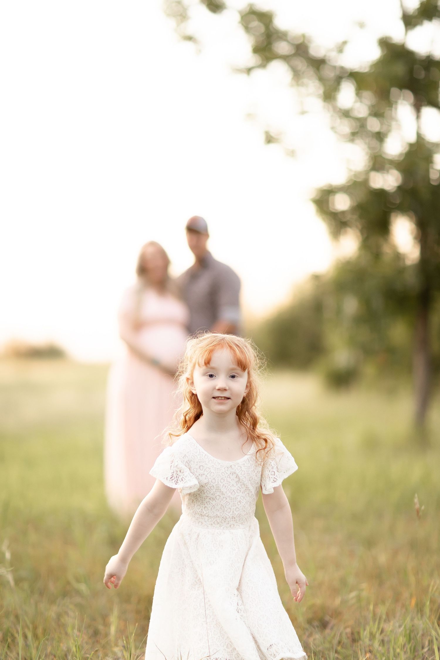 Young redhead girl in white dress walks toward camera with expecting parents softly blurred in background.