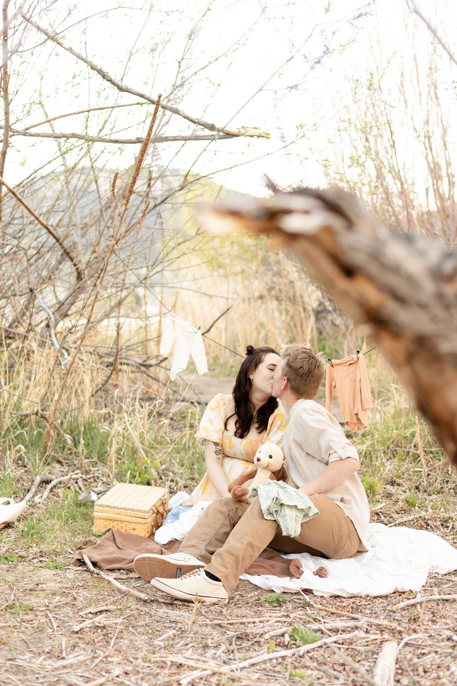 Expecting couple shares a kiss while sitting on blanket with baby items and clothesline of baby clothes behind them.