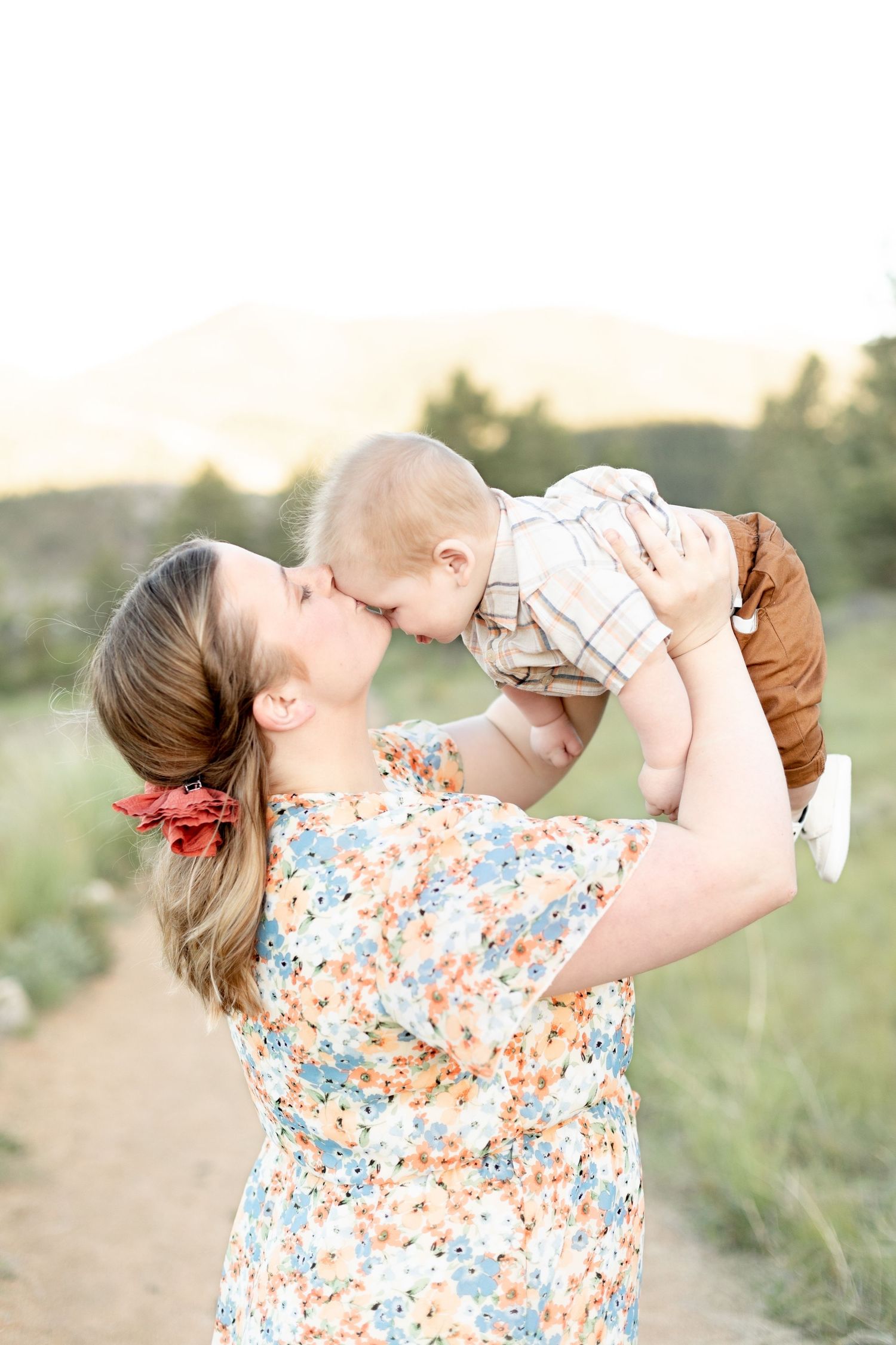 Mother lifting baby up for a nose kiss on a sunny outdoor path in a floral dress.