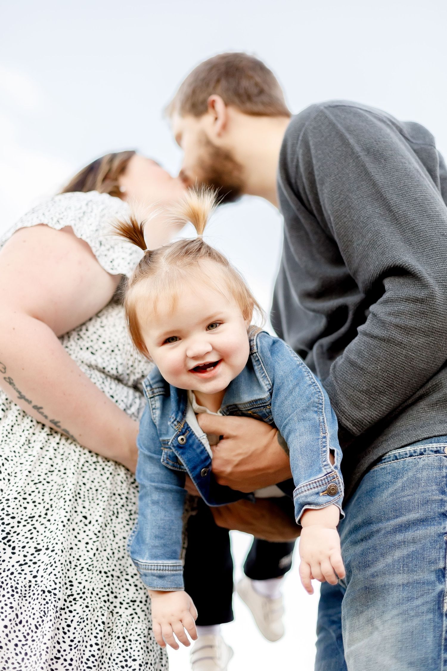 Happy toddler girl in denim jacket smiles as her parents share a kiss above her outdoors.