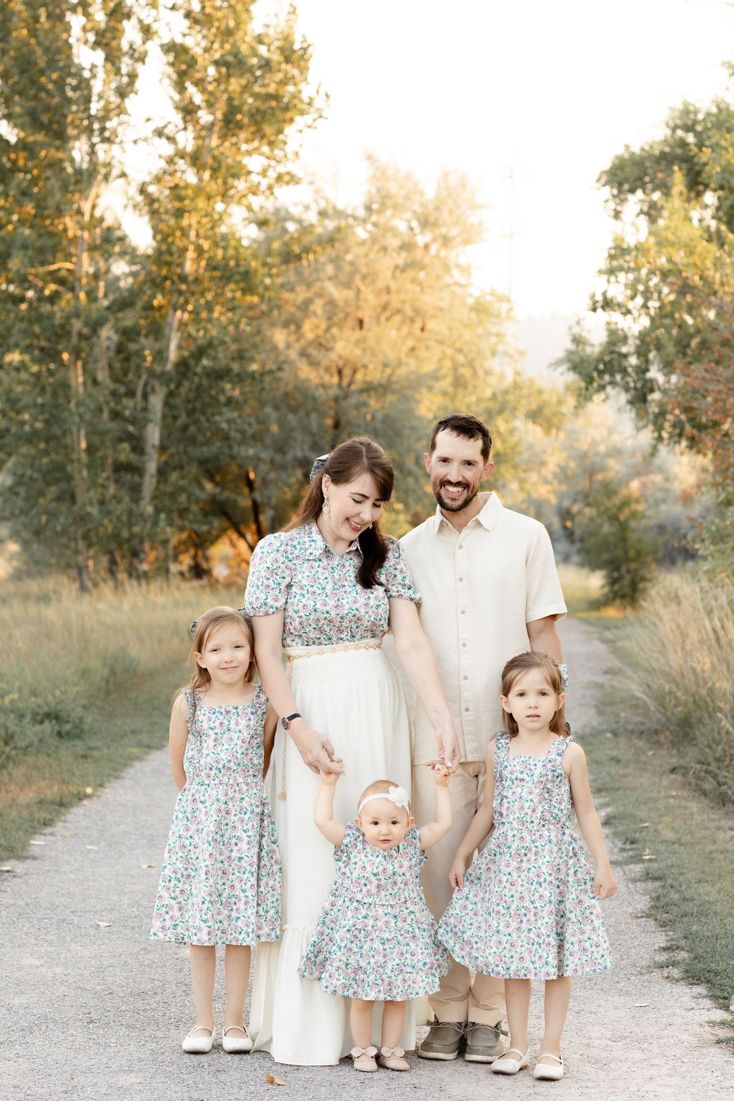 A smiling family with three young girls in matching floral dresses pose on a sunlit path.