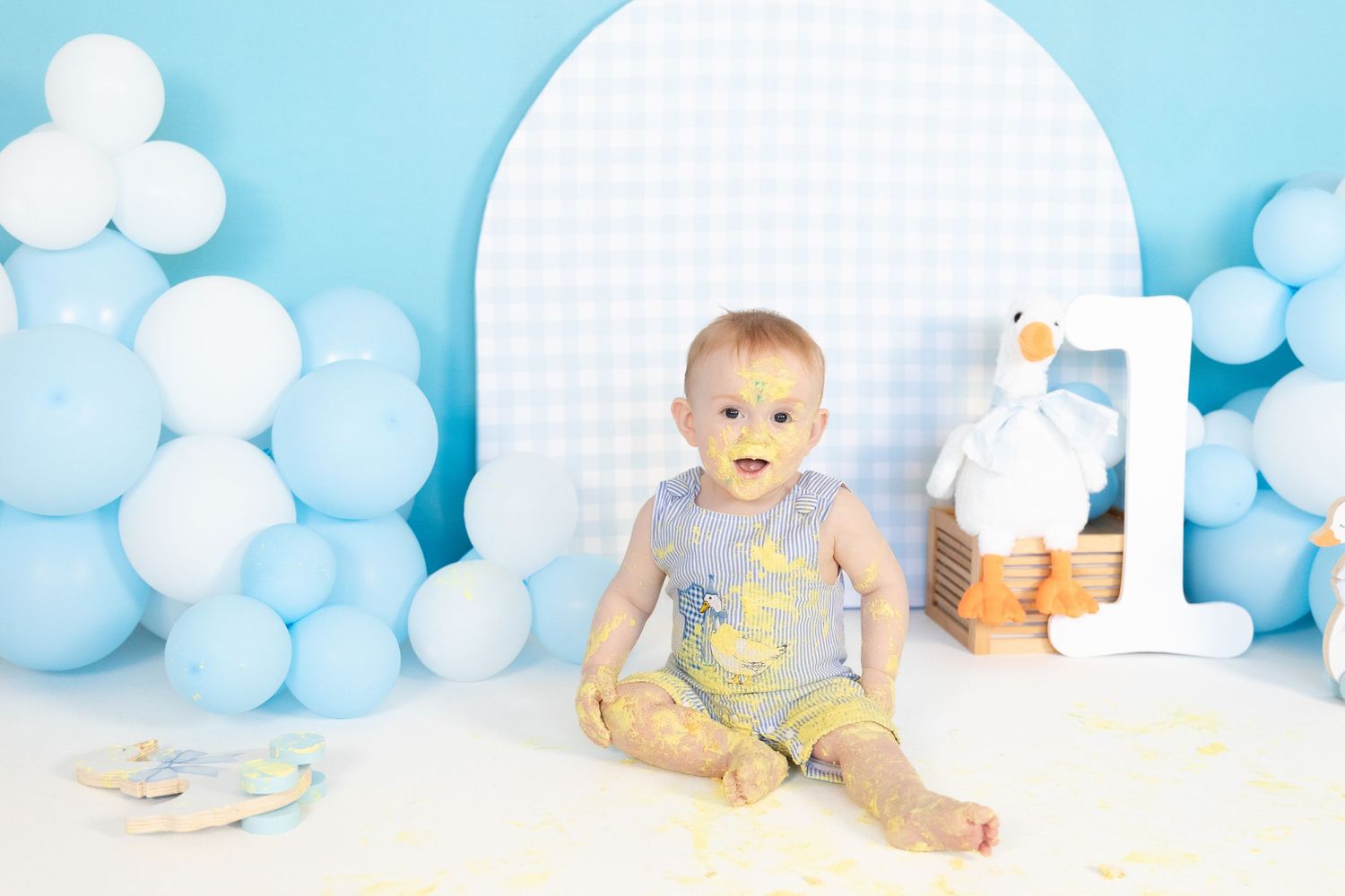 Clean baby boy sitting with blue balloons and goose decorations after first birthday cake smash.