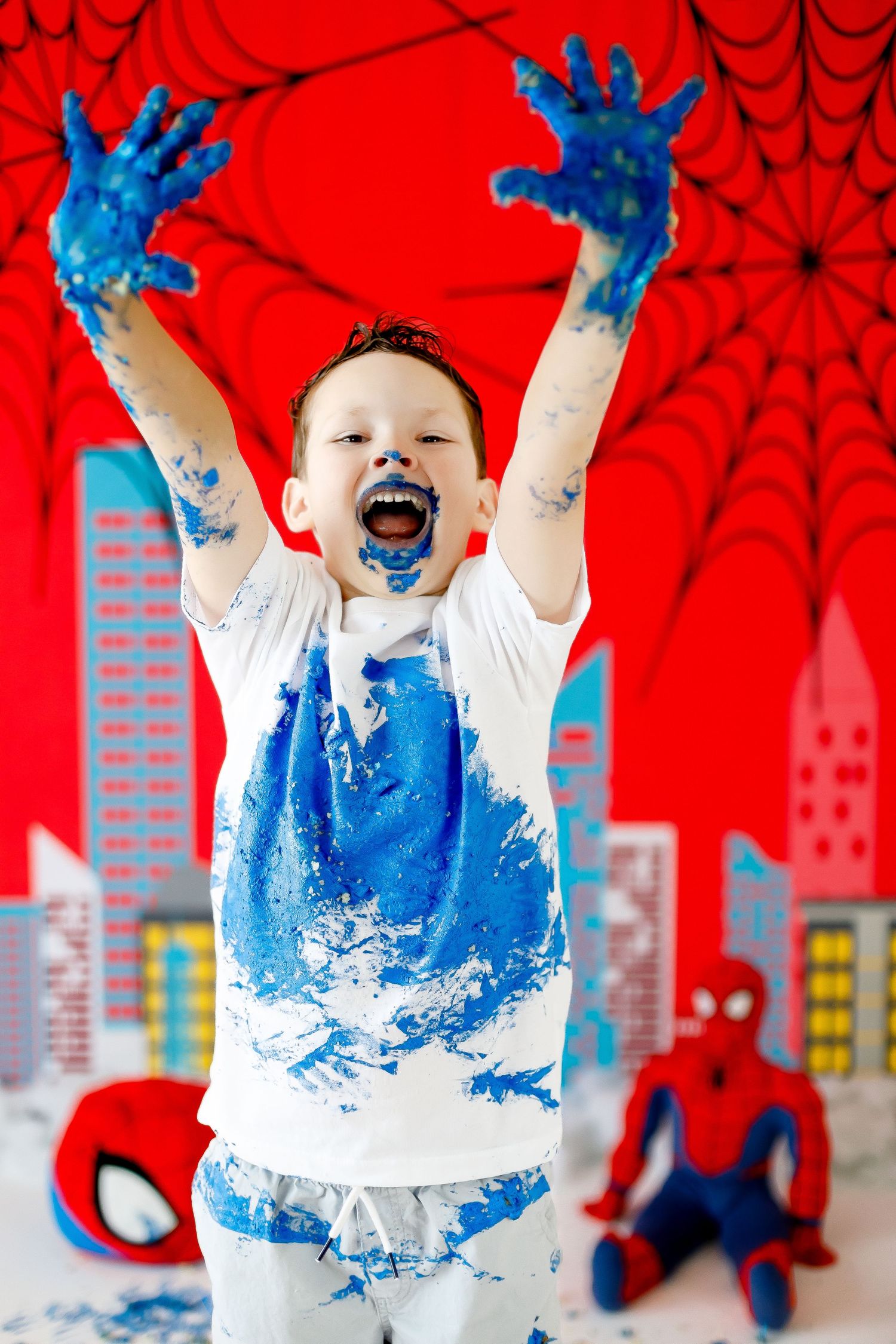 A joyful boy raises his blue frosting-covered hands at a Spiderman-themed cake smash celebration.