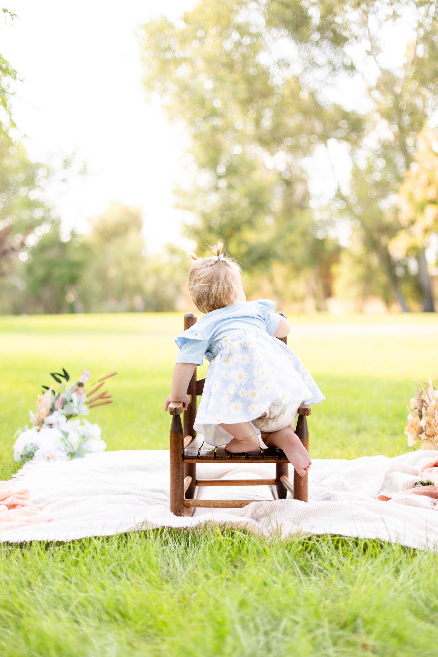 Baby girl in a blue floral dress sitting in a small wooden chair on a blanket in a sunny park.