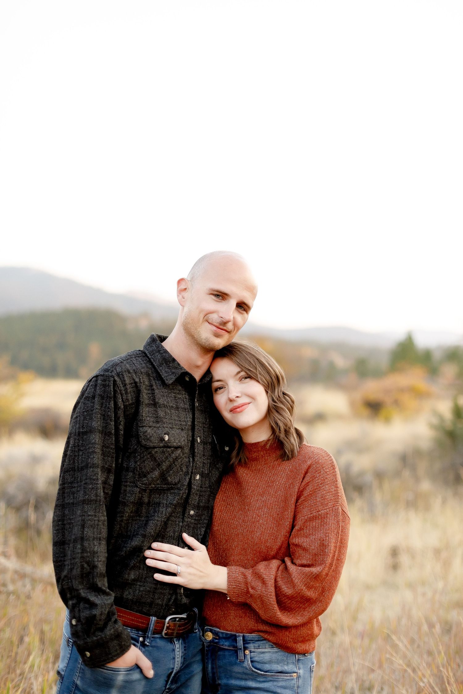 Happy couple snuggling close together in a dry autumn field with mountain landscape behind them.