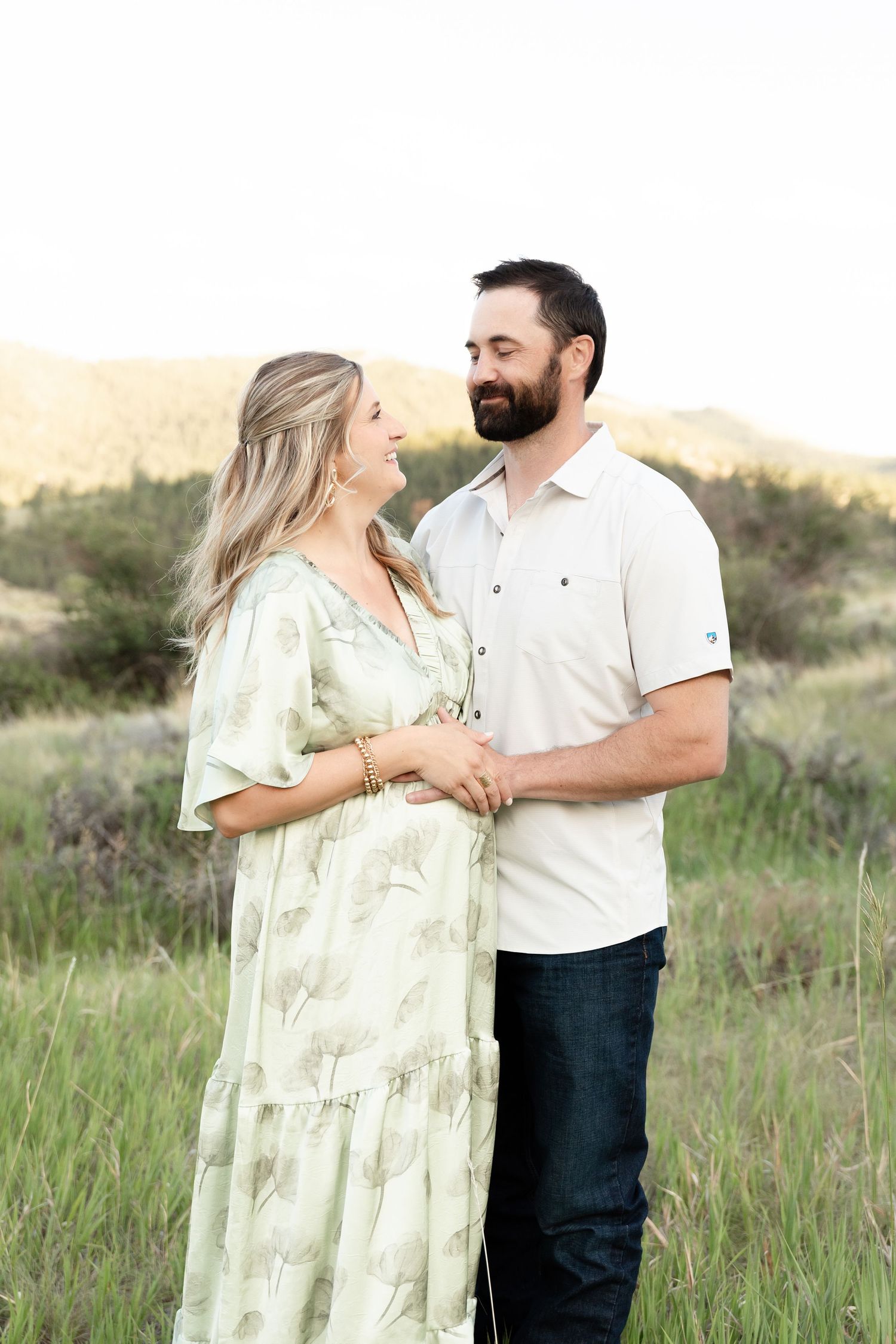 Pregnant couple holding hands and gazing at each other lovingly in a sunlit meadow.