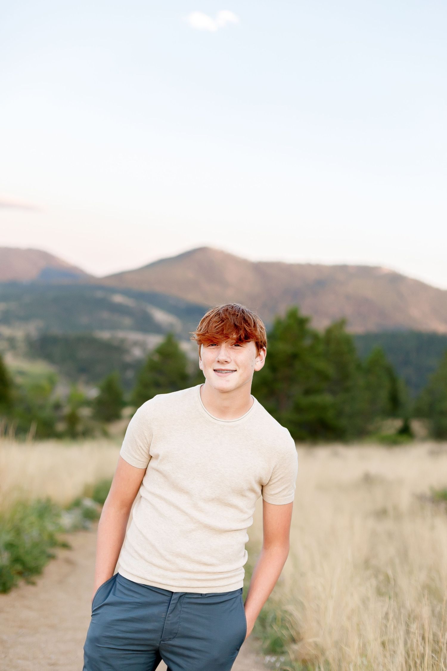 Smiling young man with red hair in beige t-shirt standing on a mountain trail at dusk.