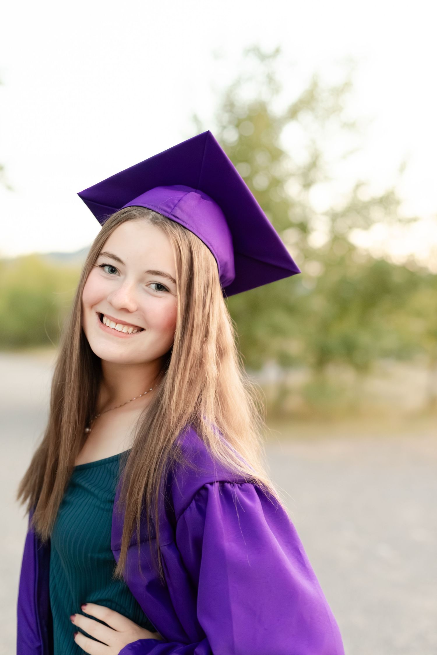 Smiling high school senior in purple graduation cap and gown posing outdoors in warm evening light.